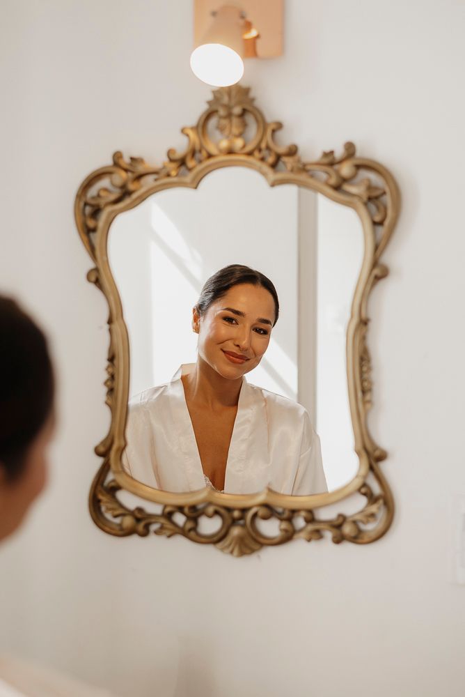 A series of ornate gold-framed mirrors on a white wall reflect someone wearing a white robe in warm lighting.