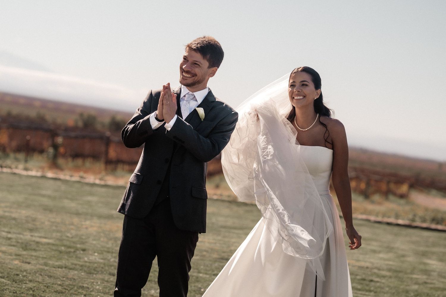 Smiling bride in white gown and veil walks with groom in black suit outdoors at a sunny wedding venue.