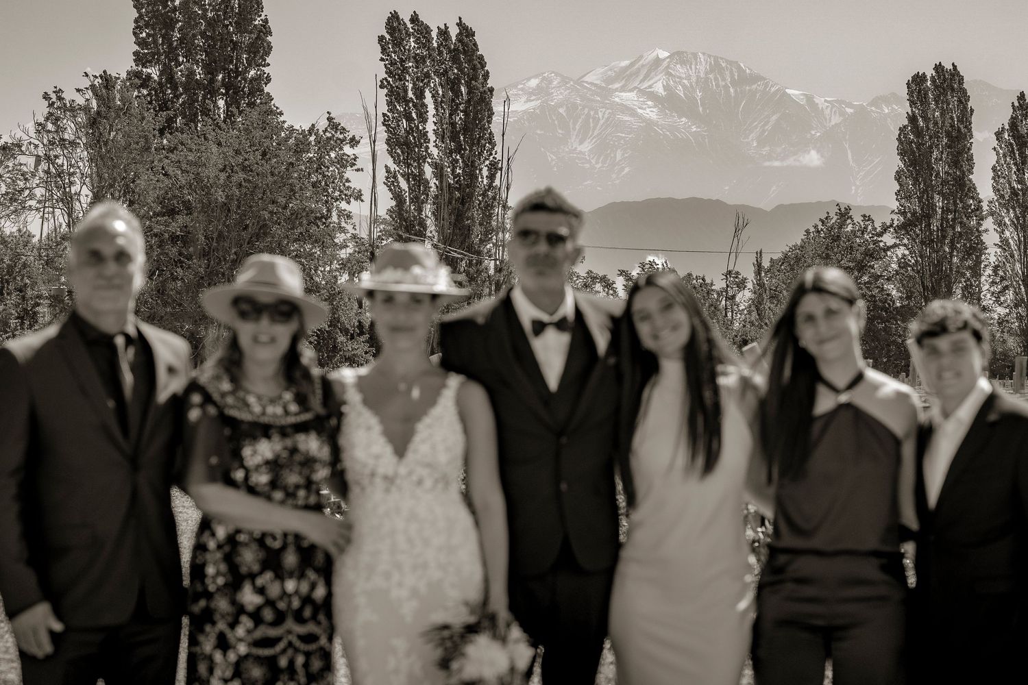 A group poses together outdoors with tall poplar trees and snow-capped mountains visible in the background.