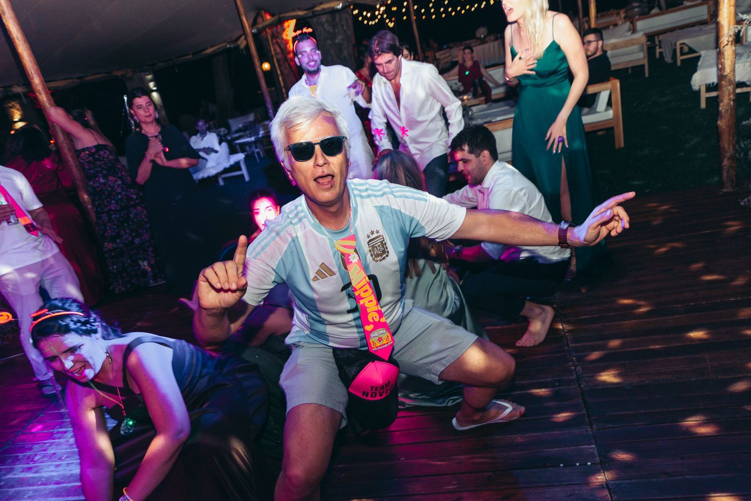 Party guests dance energetically on a wooden dance floor illuminated by colorful lights at an outdoor celebration.