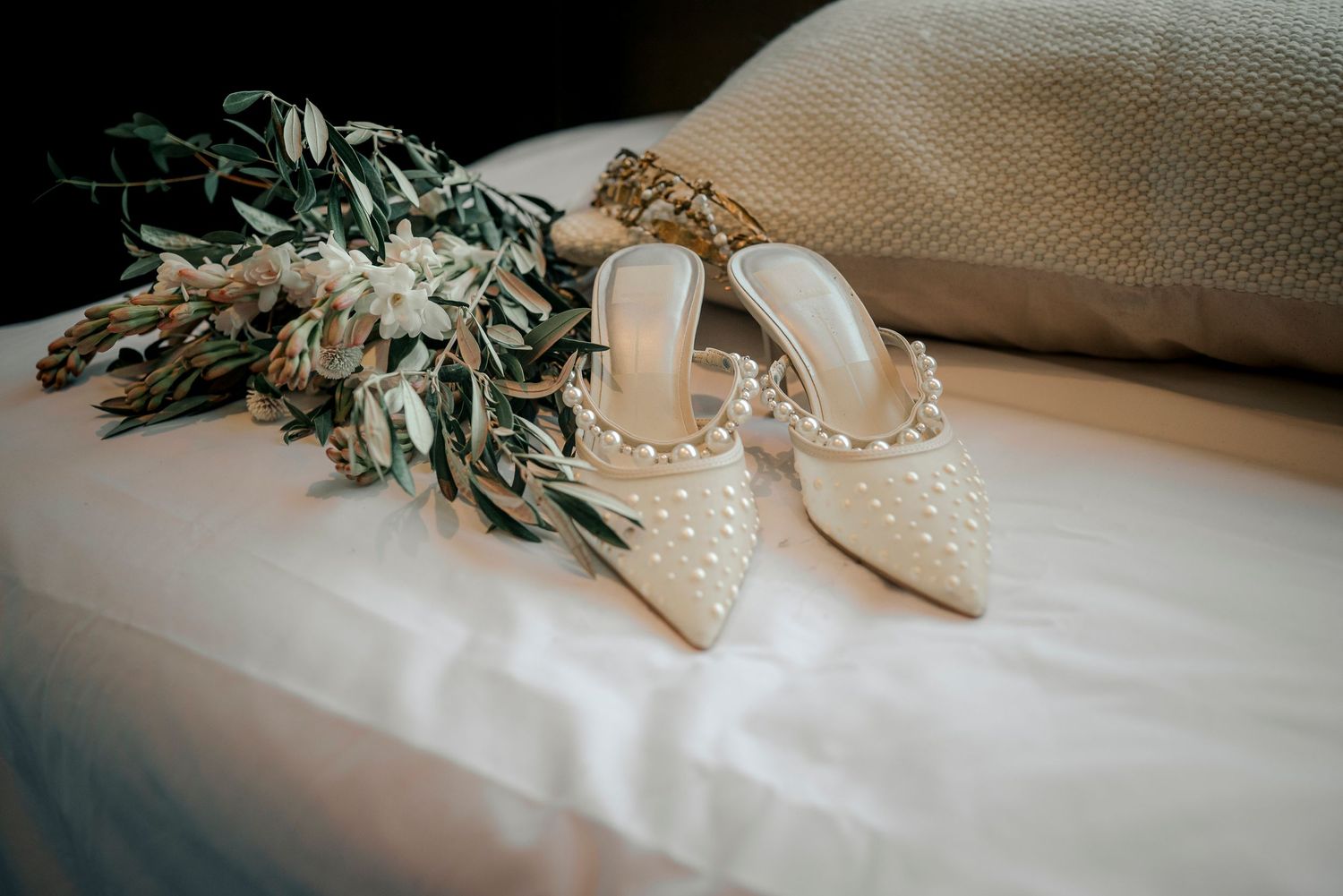 Elegant pearl-studded bridal heels displayed next to a floral bouquet on white fabric.