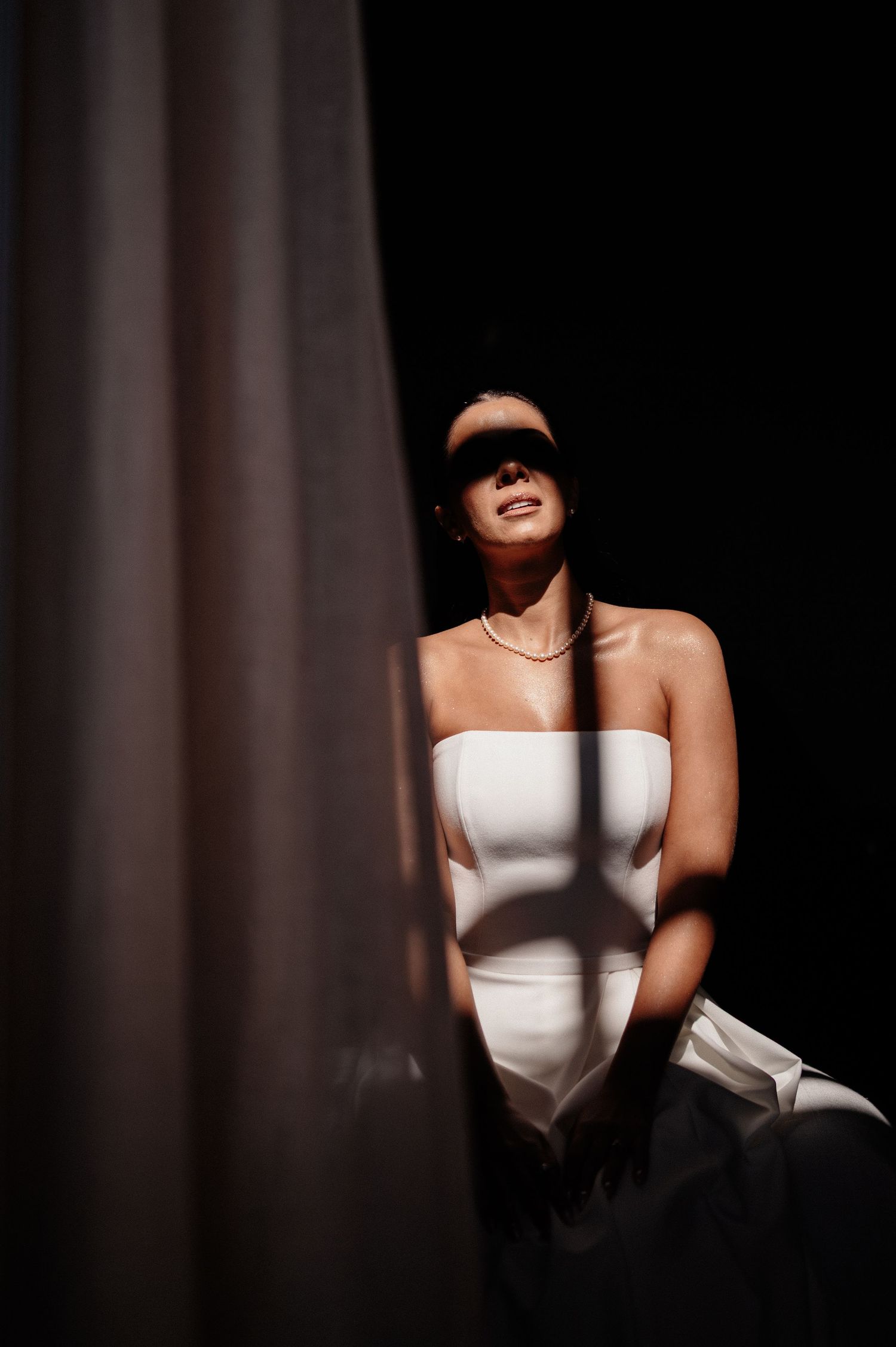 A woman in a white strapless gown stands in dramatic light and shadow, smiling with a necklace, against a dark background.