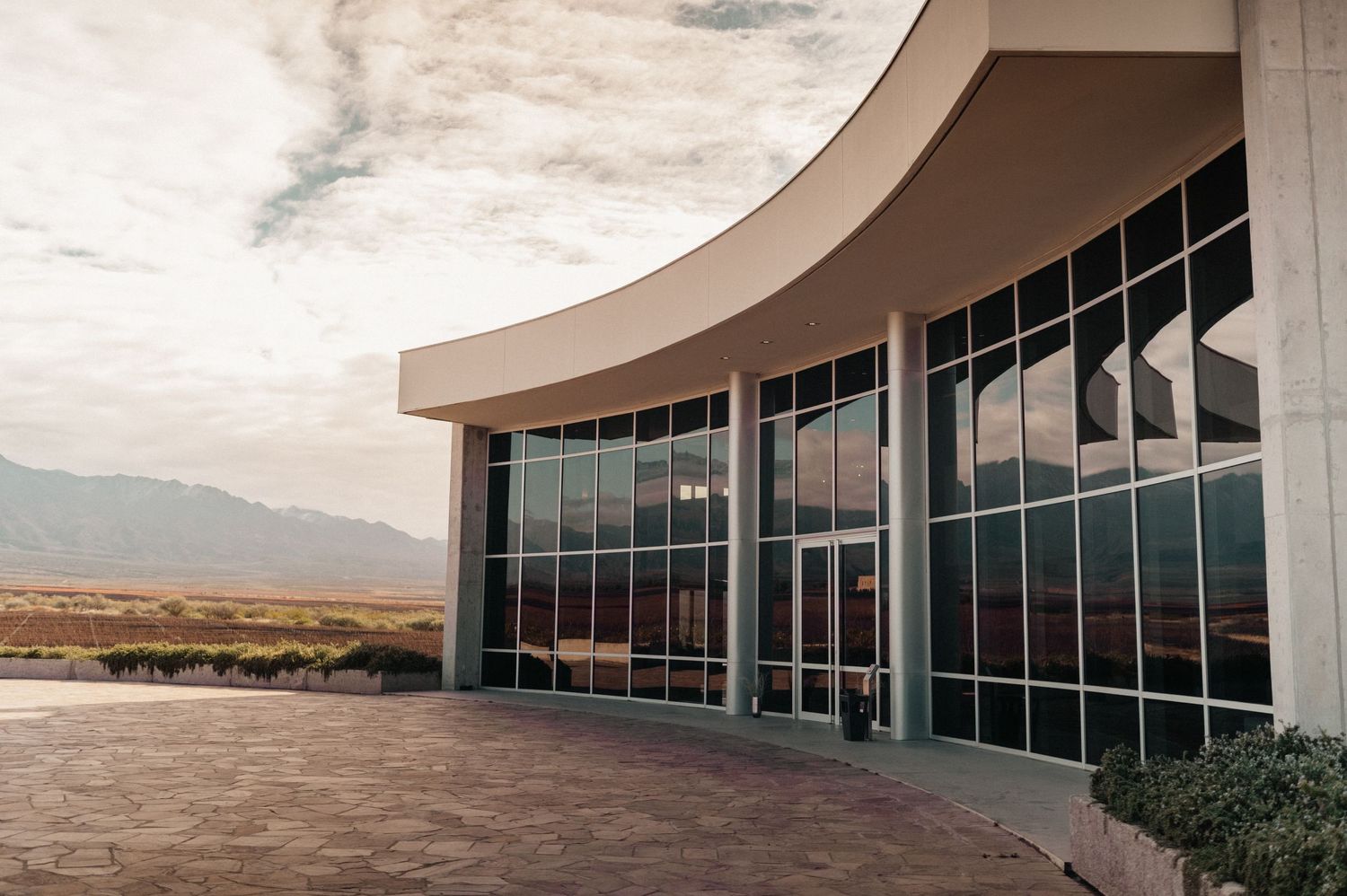 Modern curved glass building facade with mountain views and open plaza under cloudy skies.