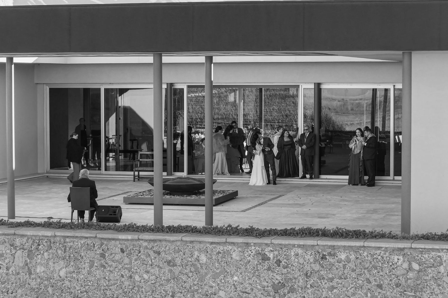 Black and white photo of a wedding ceremony outdoors under a modern pavilion with guests gathered around the couple.