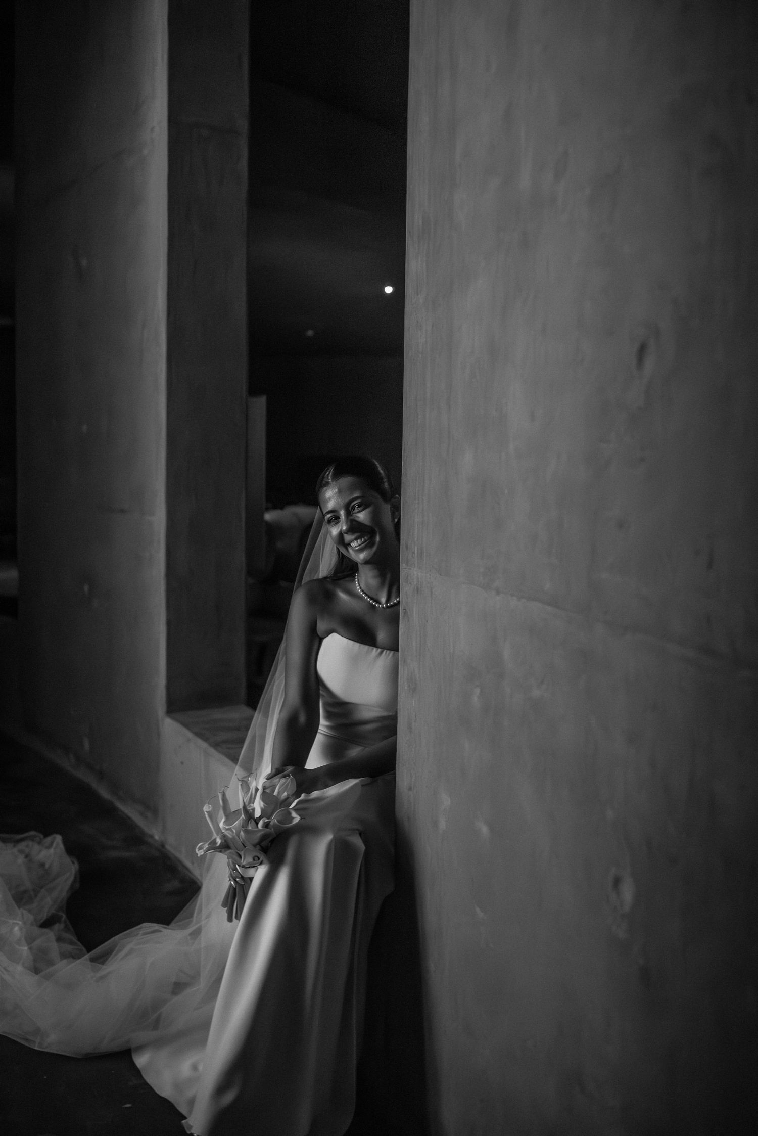 Smiling bride in strapless gown and veil poses between concrete columns in a dramatic black and white wedding portrait.