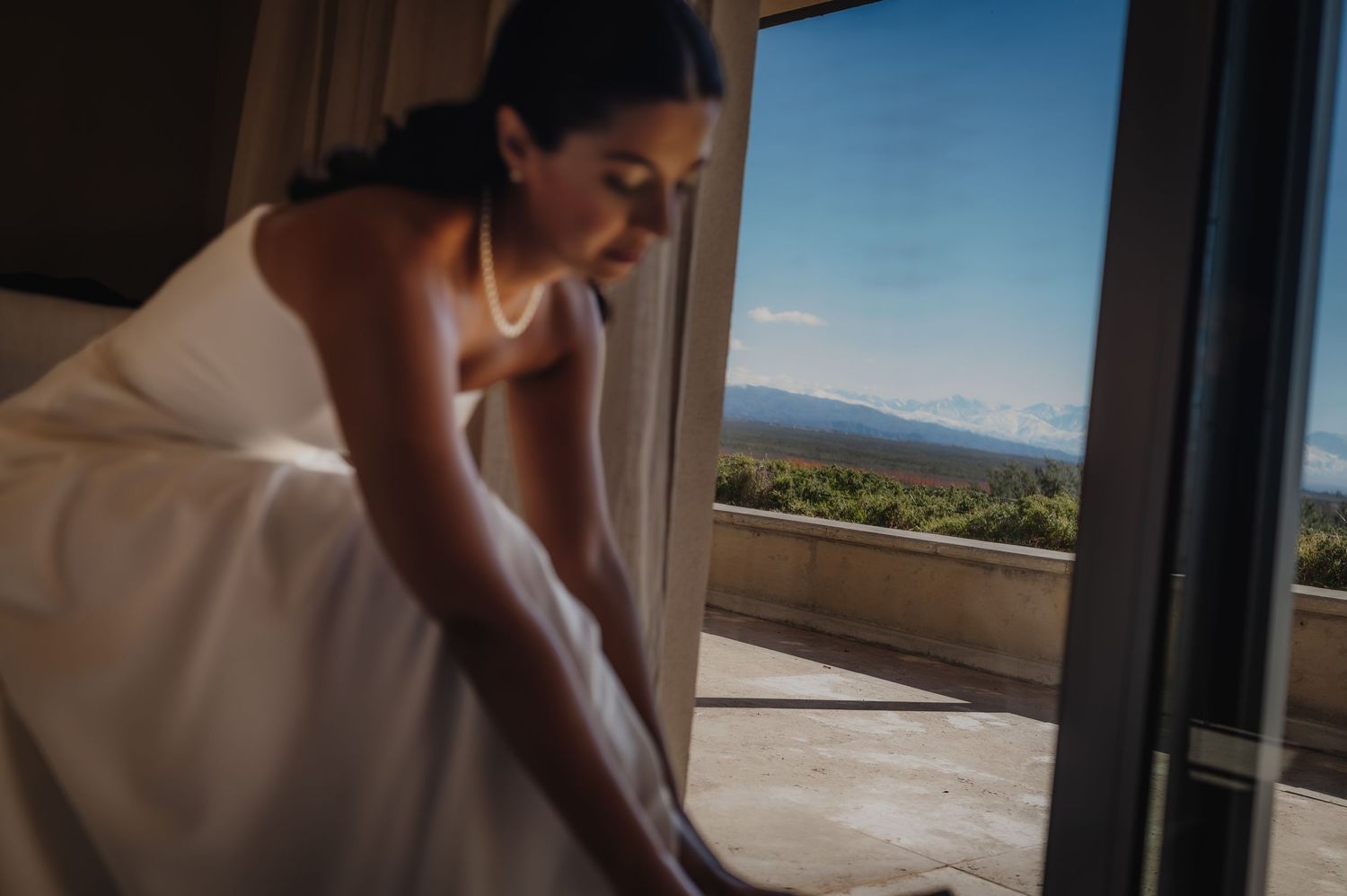 Bride in white dress adjusting her gown near an open door with scenic mountain and forest views.