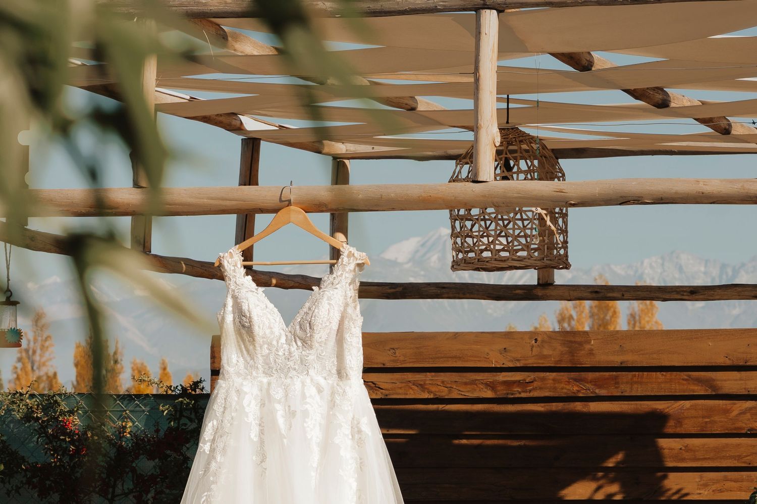 Modern wedding dress hangs from rustic wood beams at an outdoor venue with natural lighting and mountain backdrop.