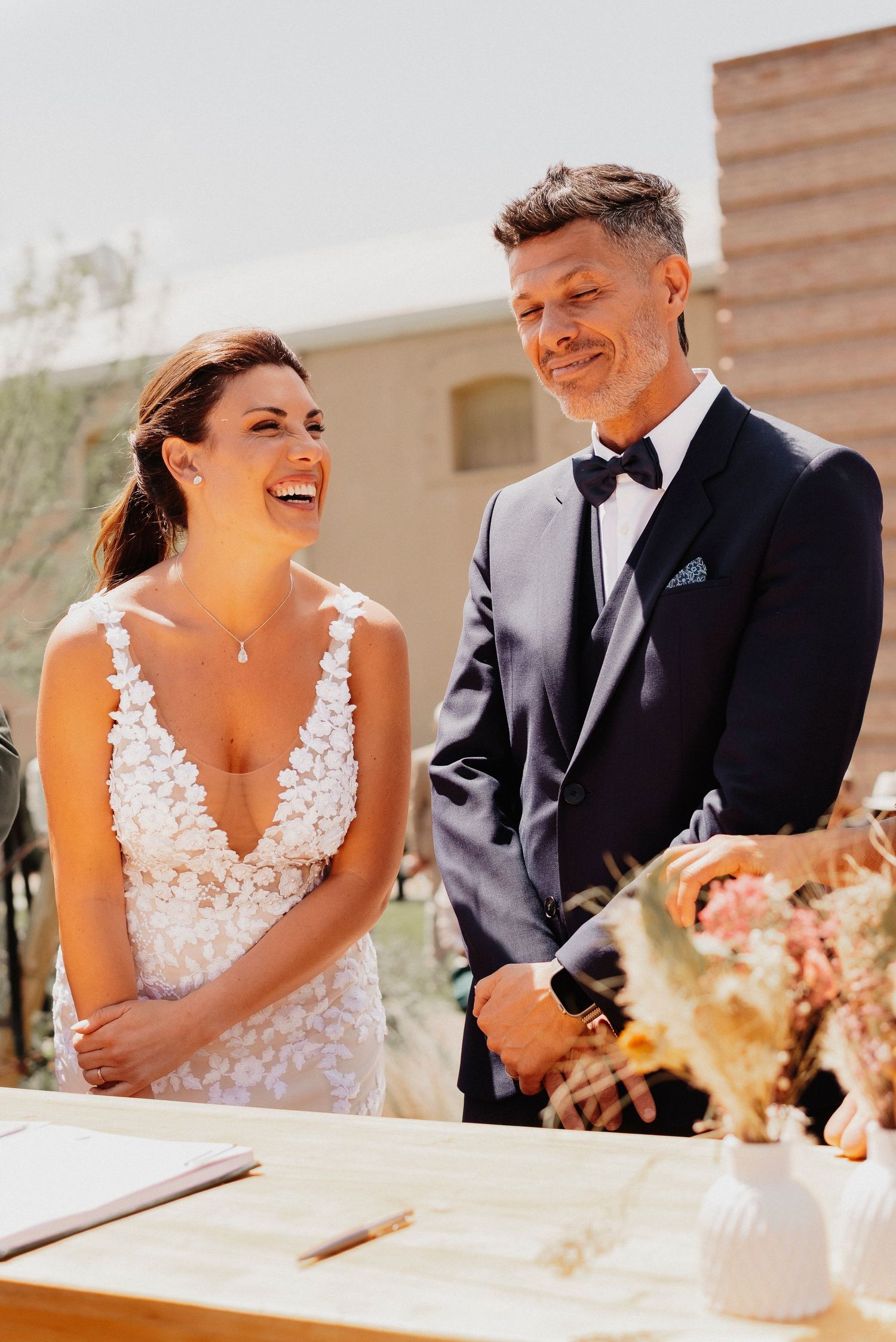 A couple shares joyful laughter during their wedding reception at an outdoor venue in warm sunlight.