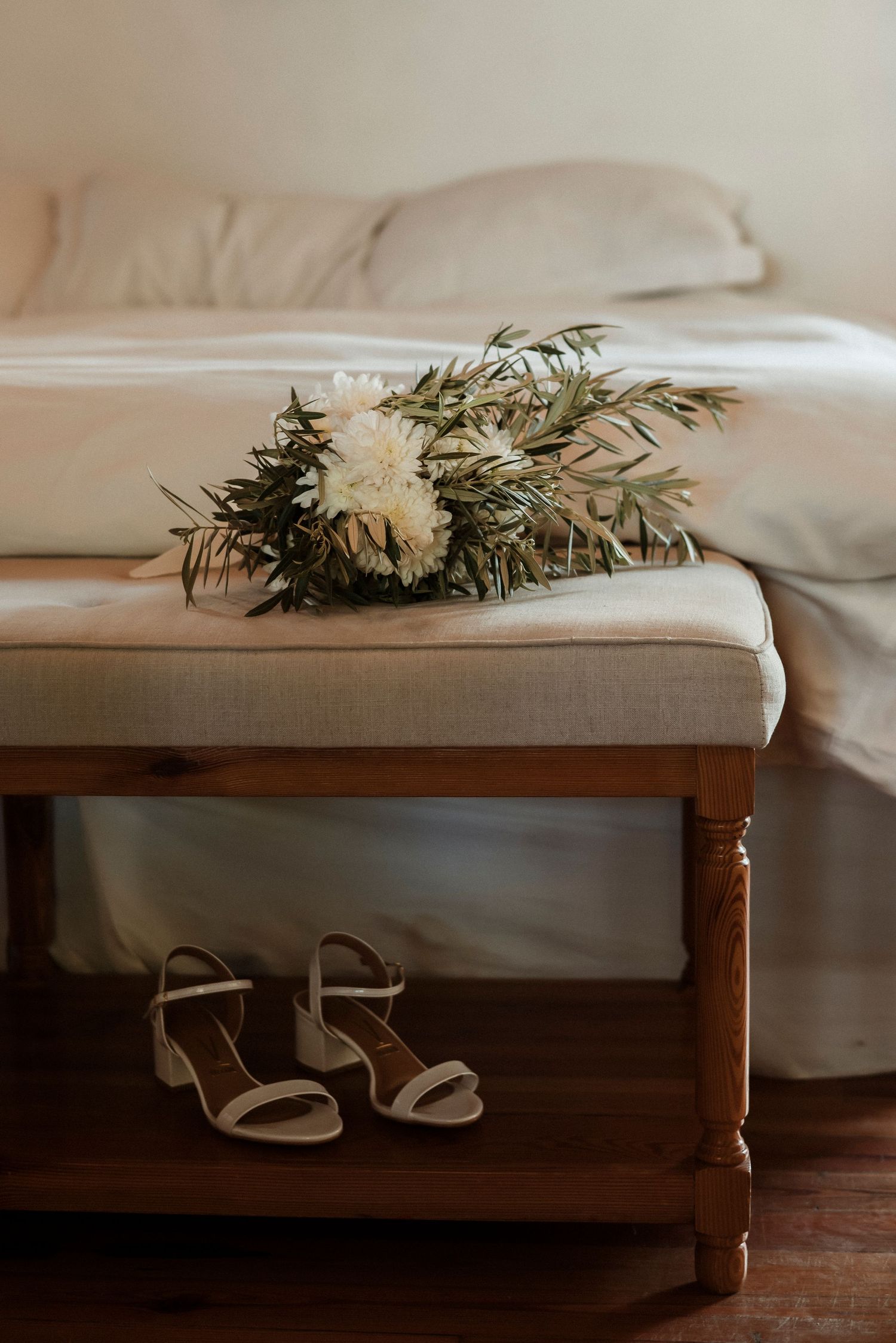 White slippers peek out beneath a bed frame with cream linens and delicate floral arrangements on top.