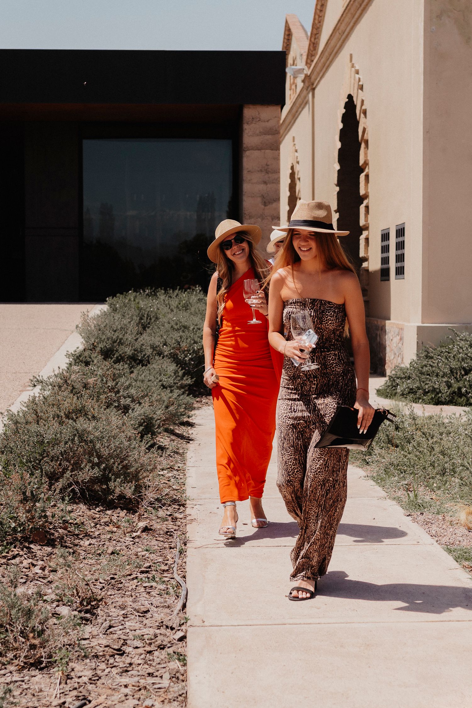 Two people walking on a concrete path next to desert landscaping wearing orange and patterned summer dresses.