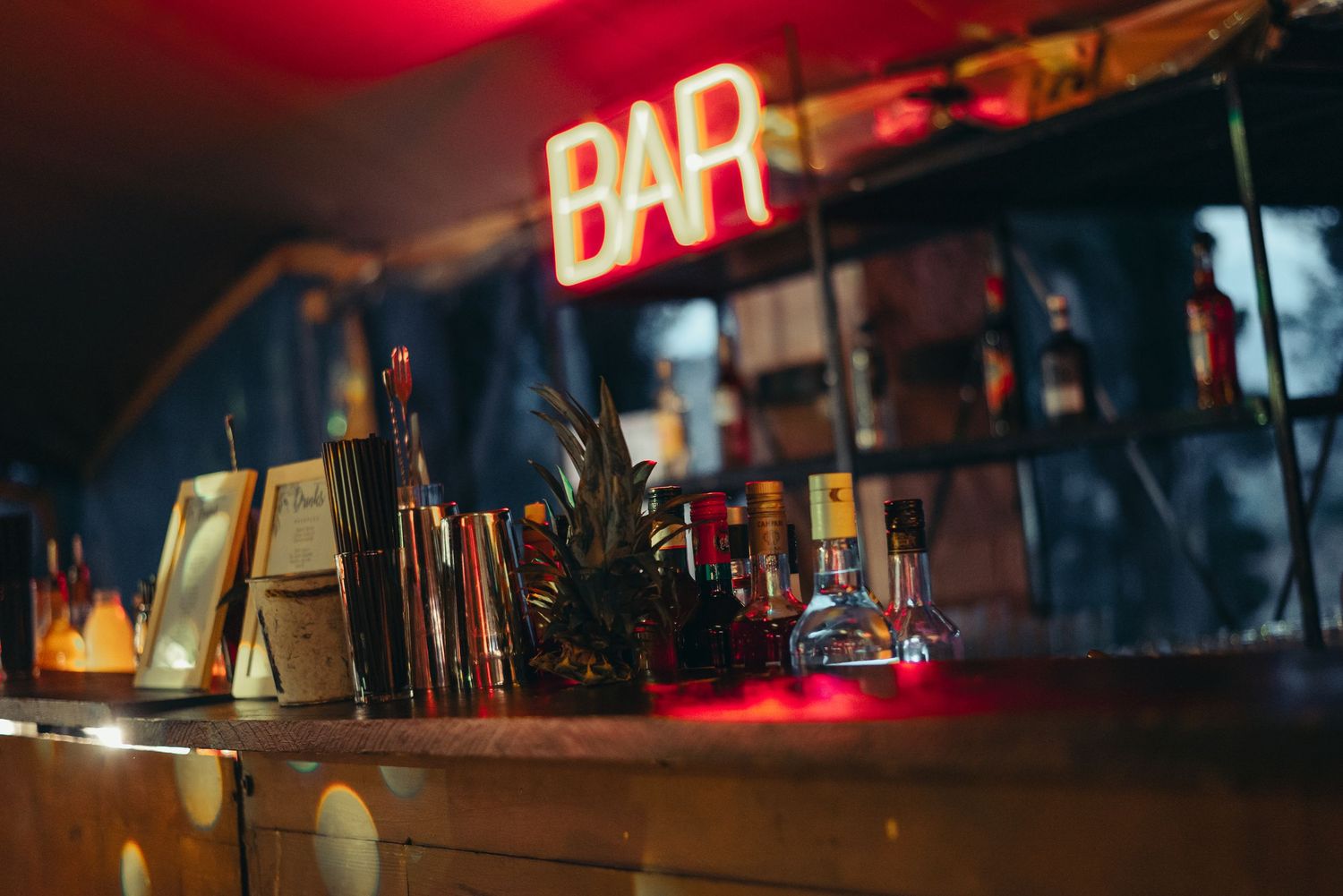 Neon bar signs illuminate a dark interior with bottles and glassware silhouetted against warm ambient lighting.