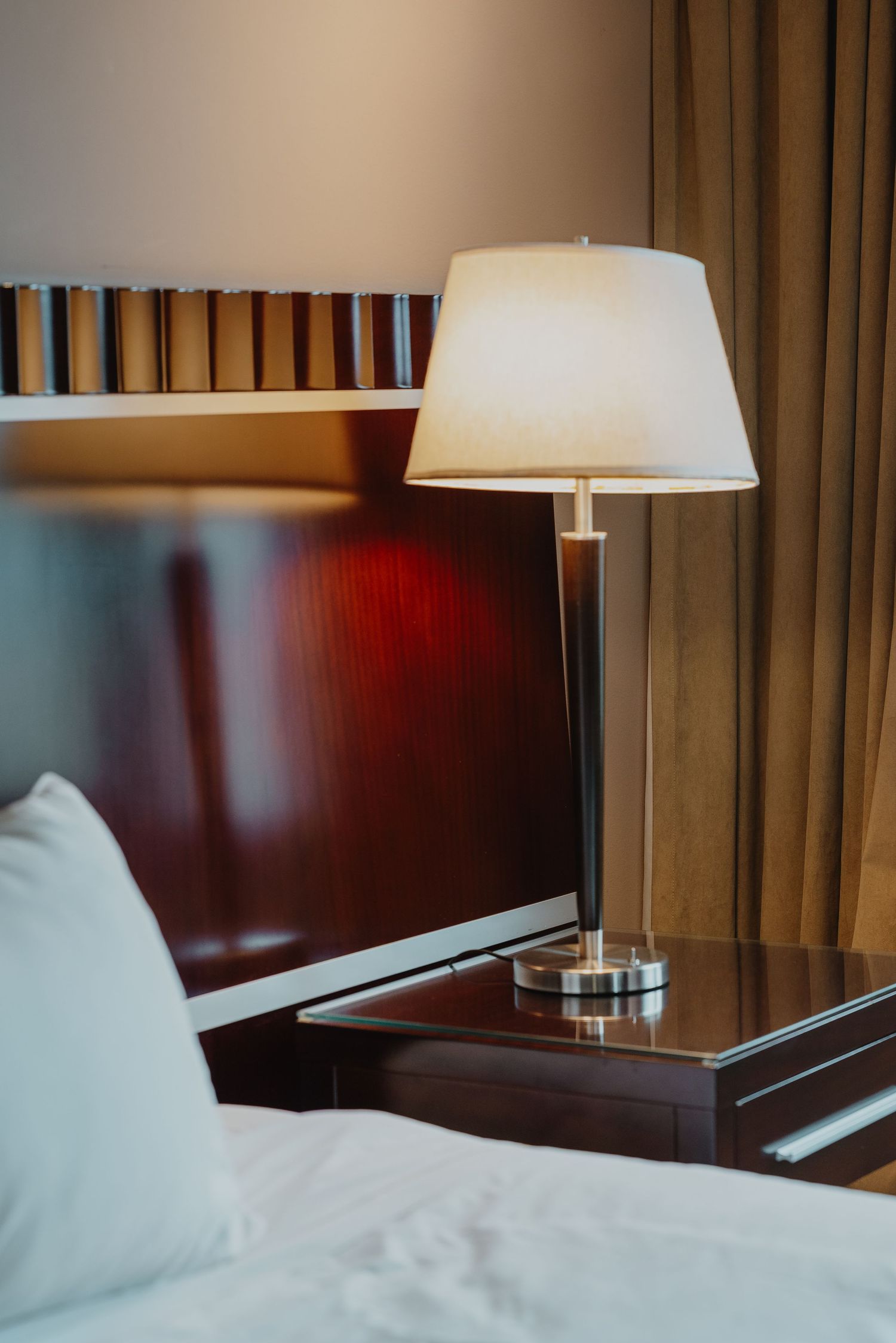 Elegant hotel bedside table with a glowing white lamp on a wooden nightstand next to a bed with white pillows.