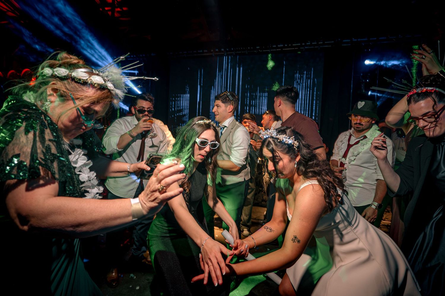 Wedding guests in floral crowns dancing energetically at a vibrant, green-lit reception party.