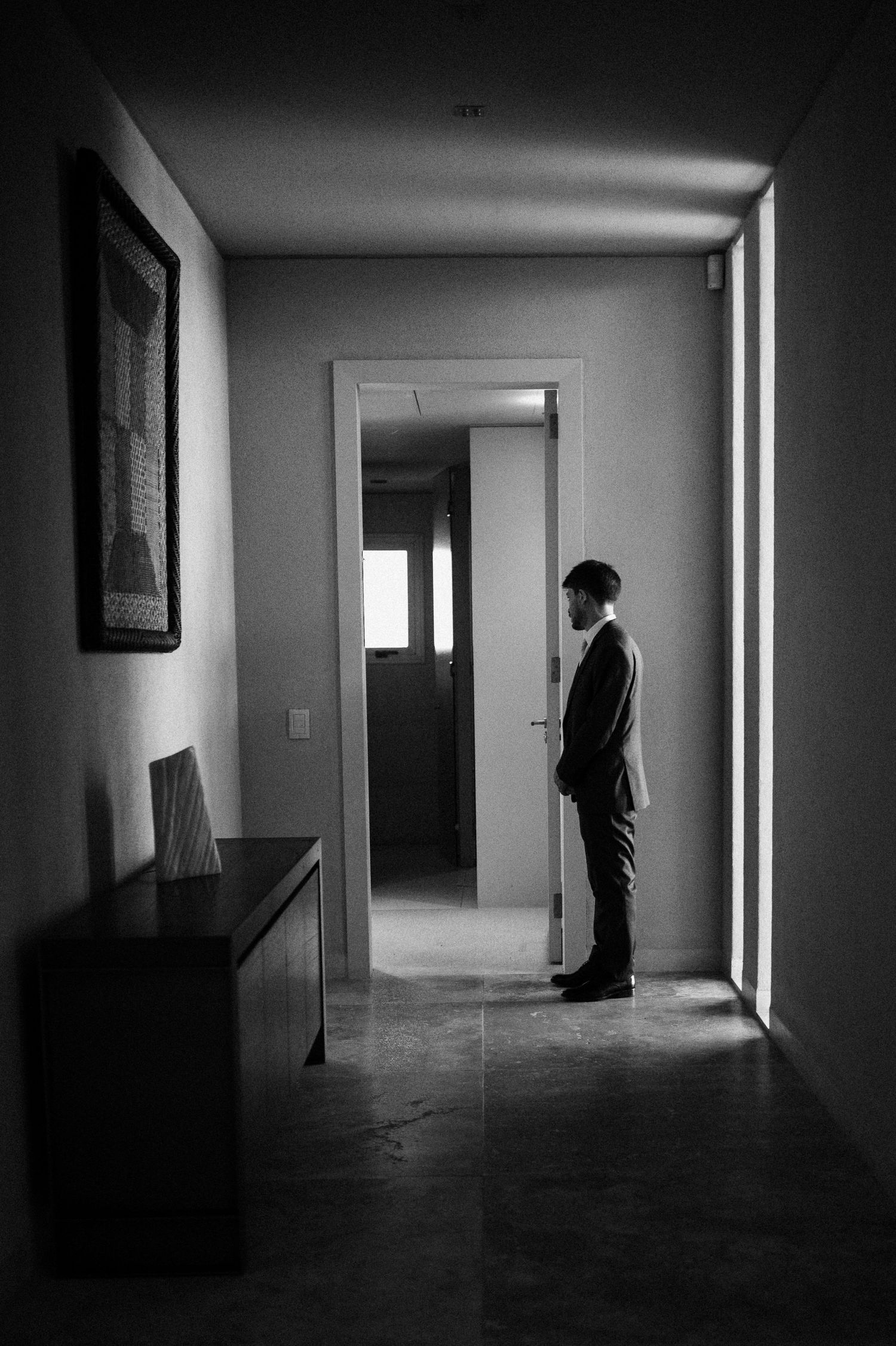 Black and white photo of a man in a suit standing in a hallway near a doorway with natural light.
