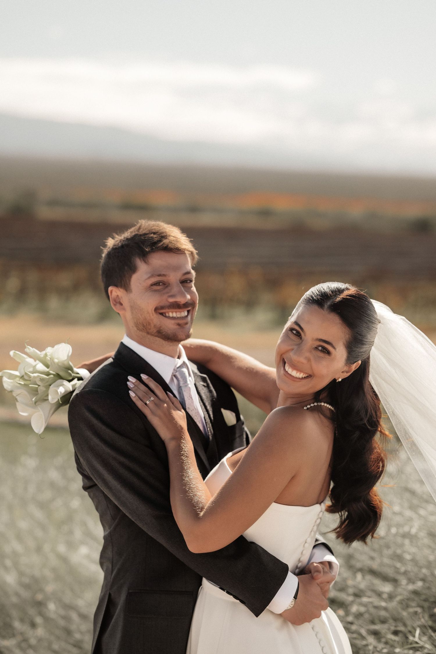 Smiling bride and groom embrace outdoors on their wedding day, with a scenic vineyard landscape in the background.