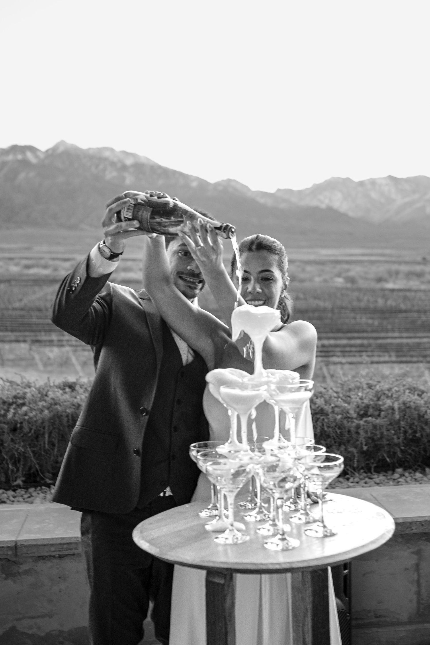 Bride and groom pouring champagne tower at outdoor wedding reception with mountain backdrop.