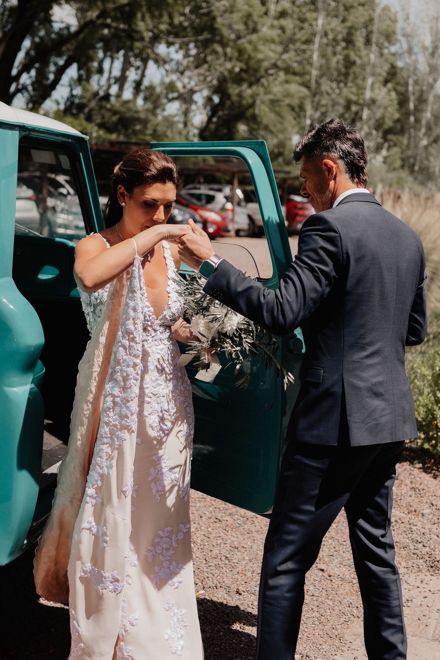 A bride in a lace gown and groom in a dark suit stand beside a vintage turquoise car during their wedding photoshoot.