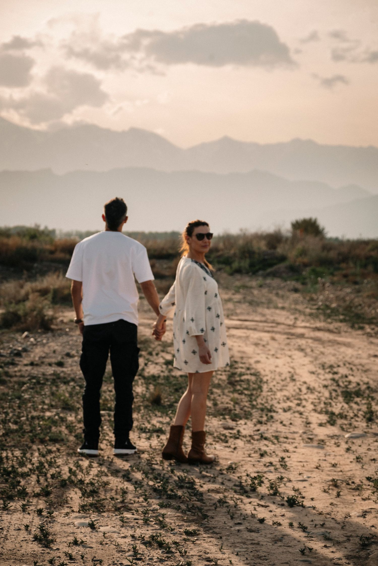 A couple walks on a dirt path at sunset with mountains and clouds creating a dreamy backdrop.