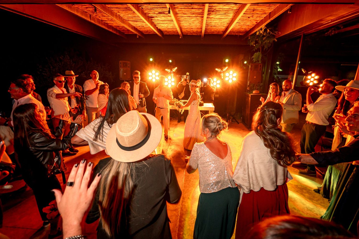 Wedding guests dancing together on a dimly lit dance floor with warm orange lighting and wooden ceiling beams.