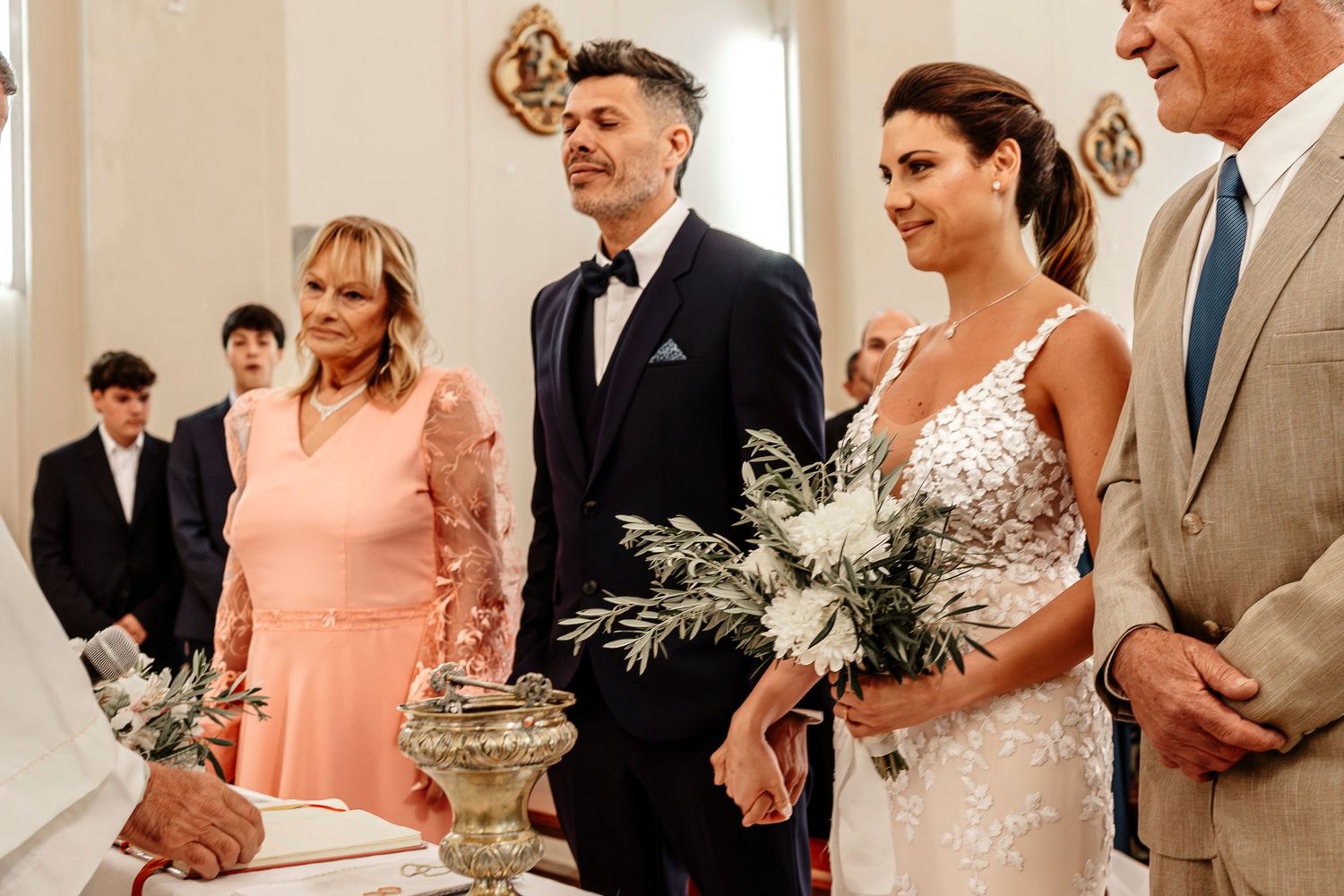 A wedding party stands in a row wearing peach and neutral colored attire during an indoor ceremony.