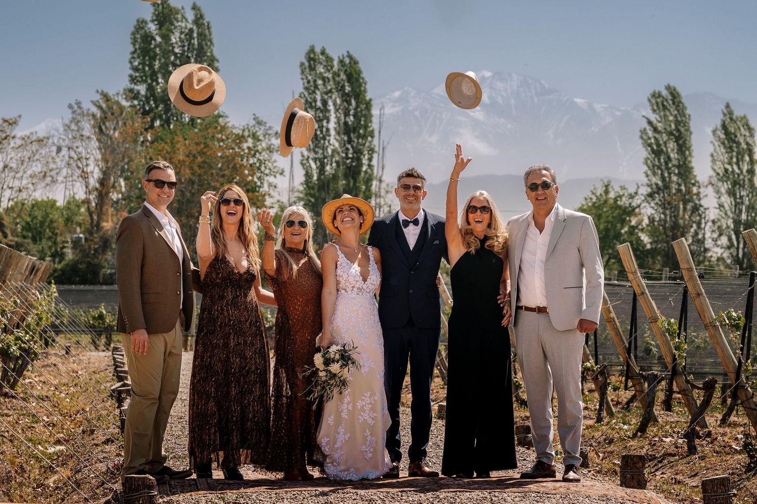 Wedding party poses outdoors with tall poplar trees in background during summer celebration.