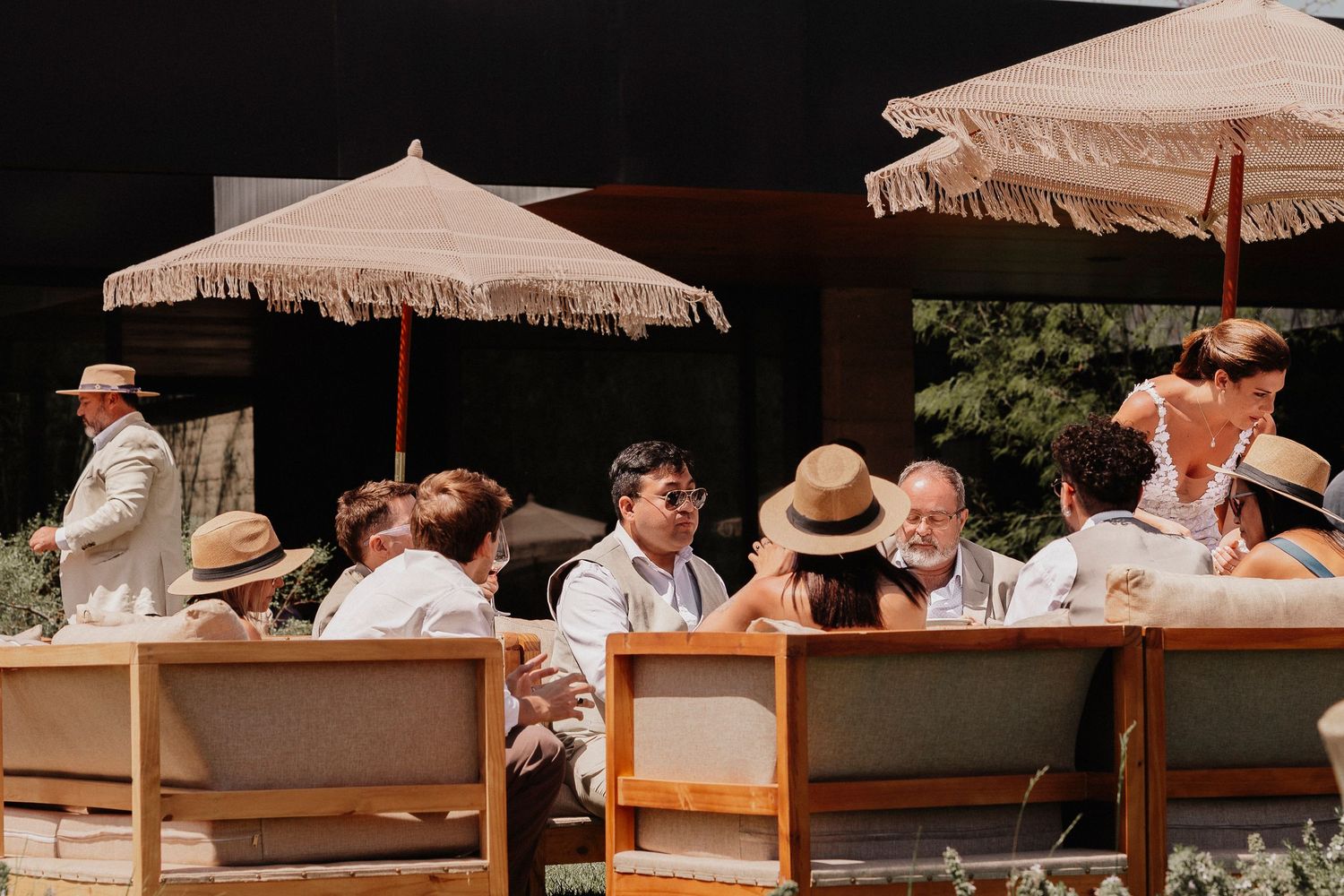 Guests relax under beige umbrellas at an outdoor event with wooden furniture on a sunny day.