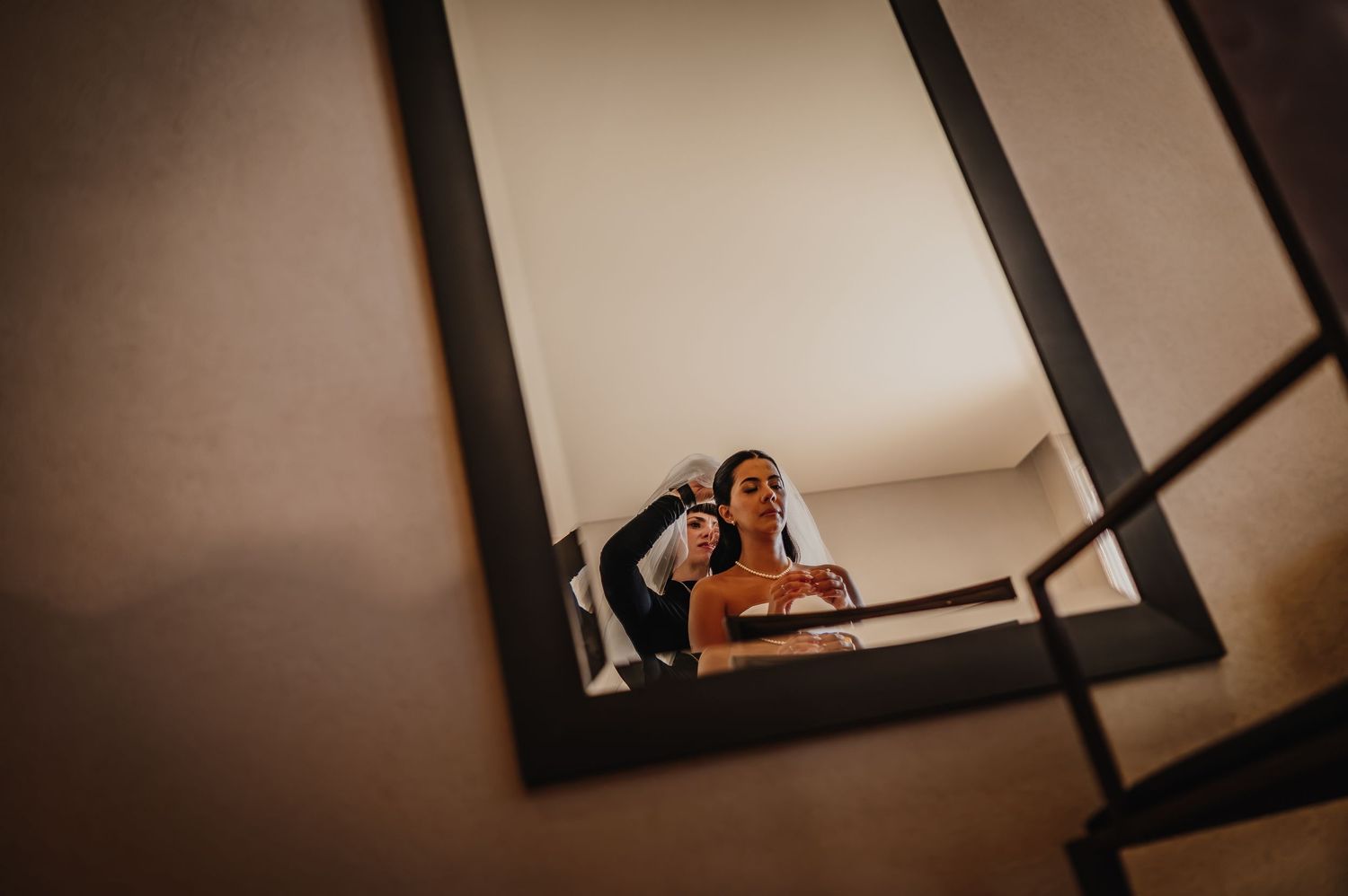 Bride getting hair styled by hairdresser, reflected in a dark-framed mirror during wedding preparations.