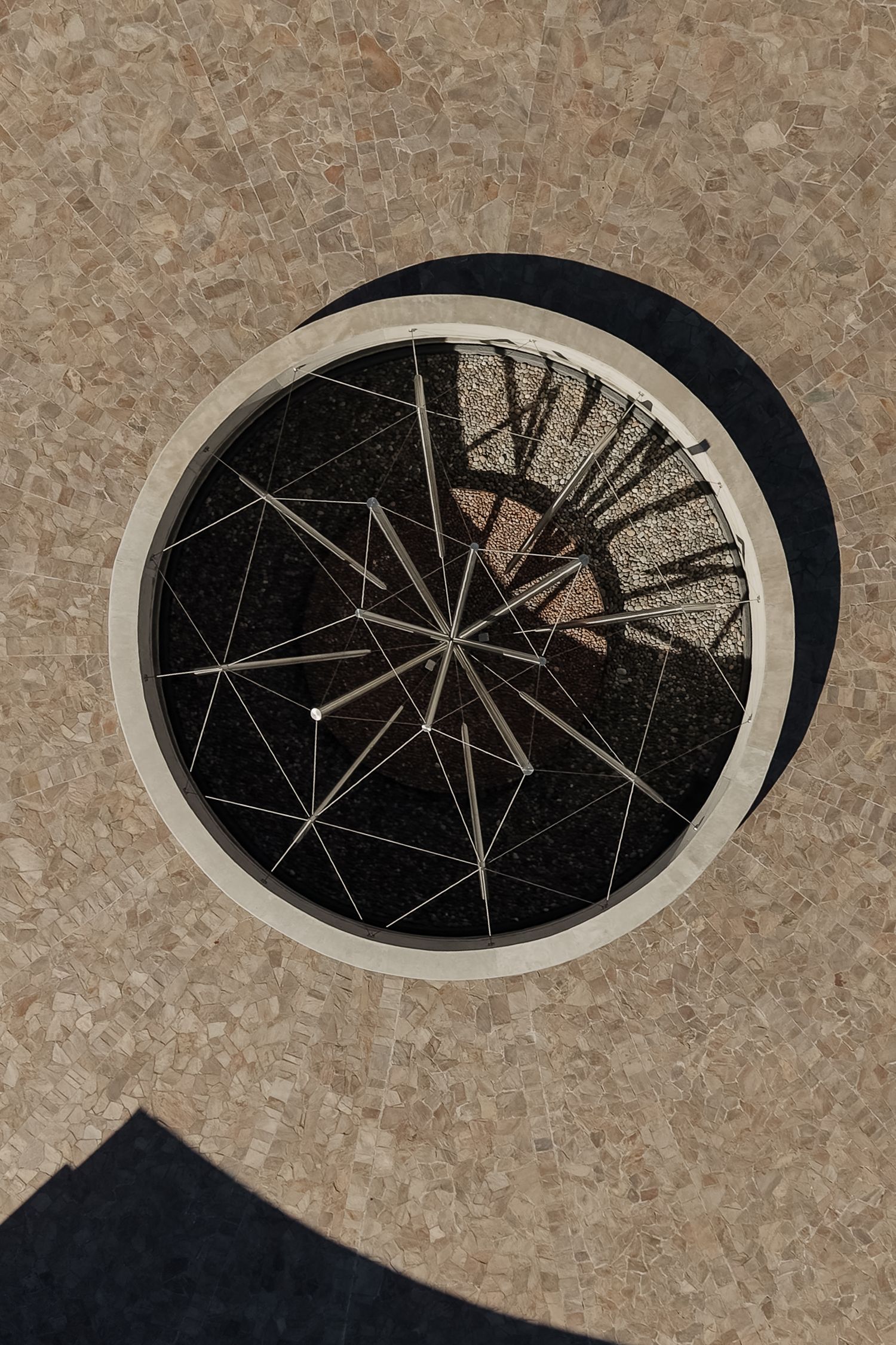 Aerial view of a circular skylight with geometric metal framework set in a textured stone floor.