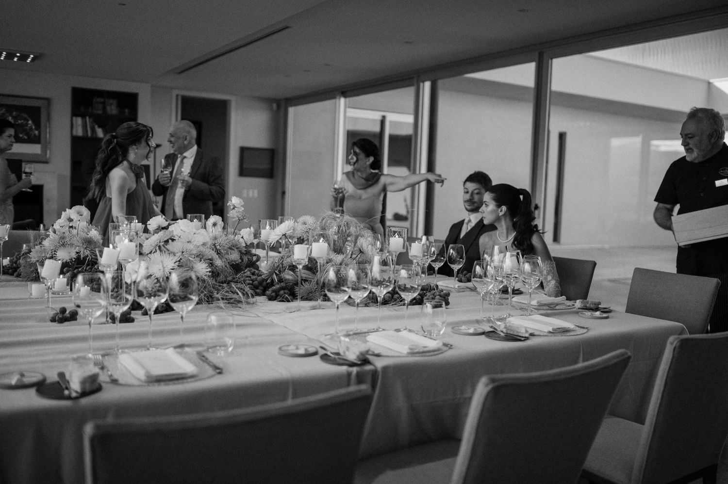 Black and white photo of wedding party gathered around elegantly decorated reception table with floral centerpieces.