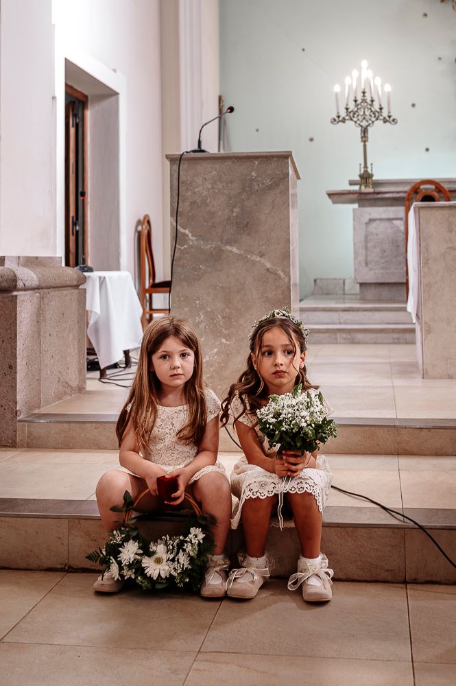 Two young flower girls in white dresses sit on stone steps with floral bouquets during a wedding ceremony. wedding in mendoza