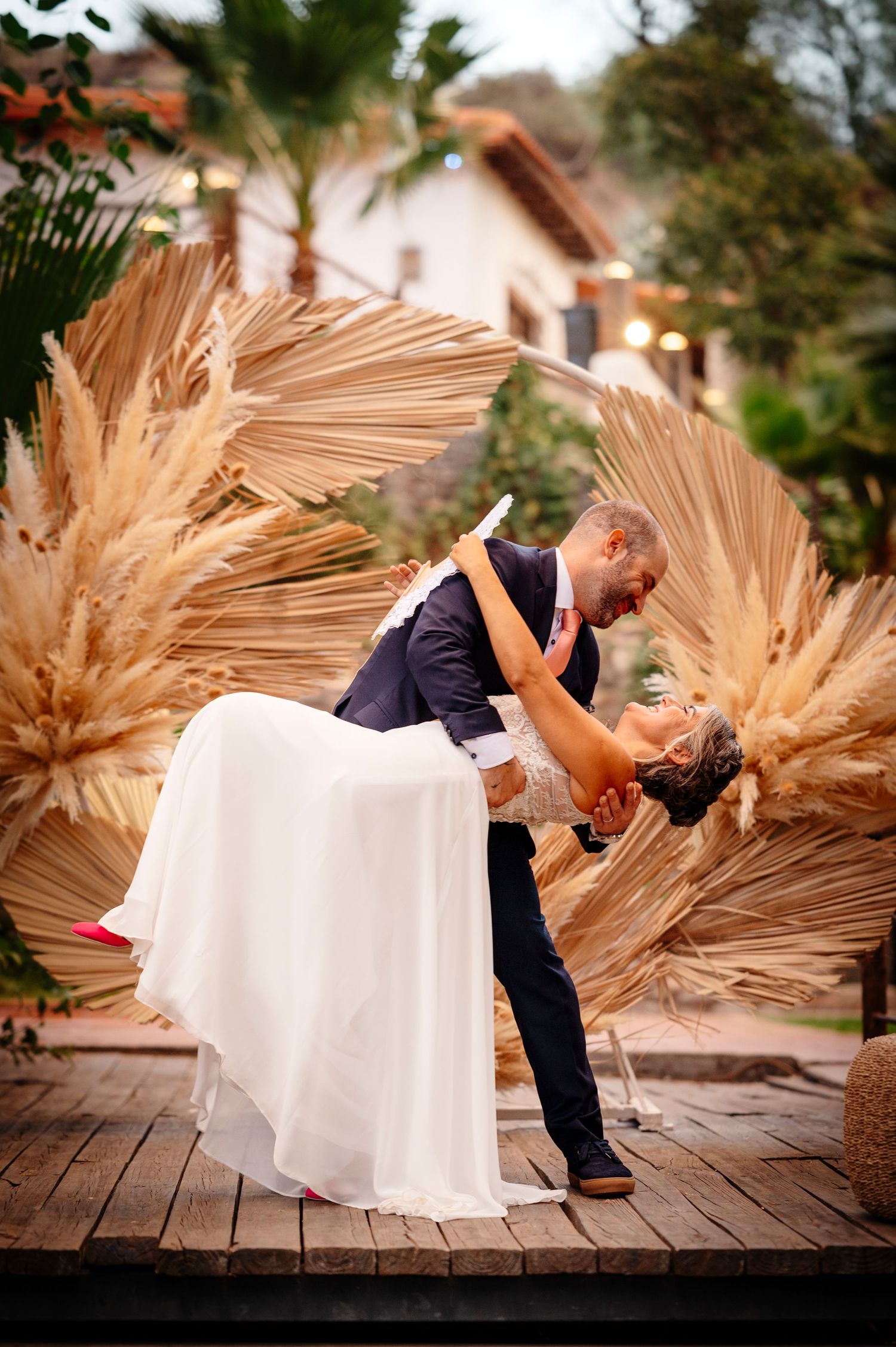 A romantic wedding dip kiss on a wooden deck surrounded by dried pampas grass arrangements at sunset.