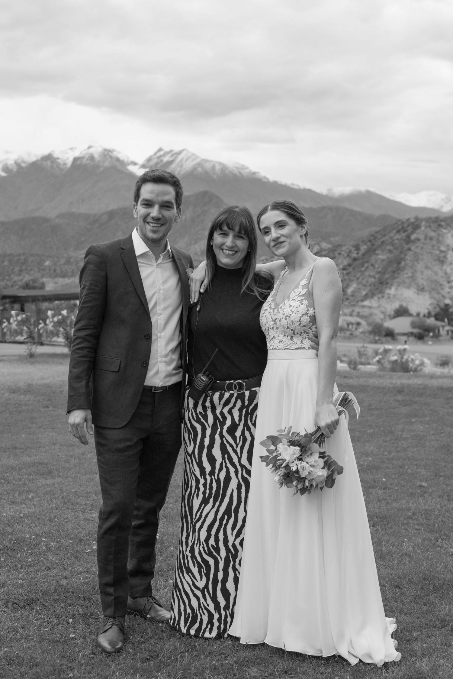A scenic wedding photo with mountain views showing three people standing together on grassy terrain in black and white.