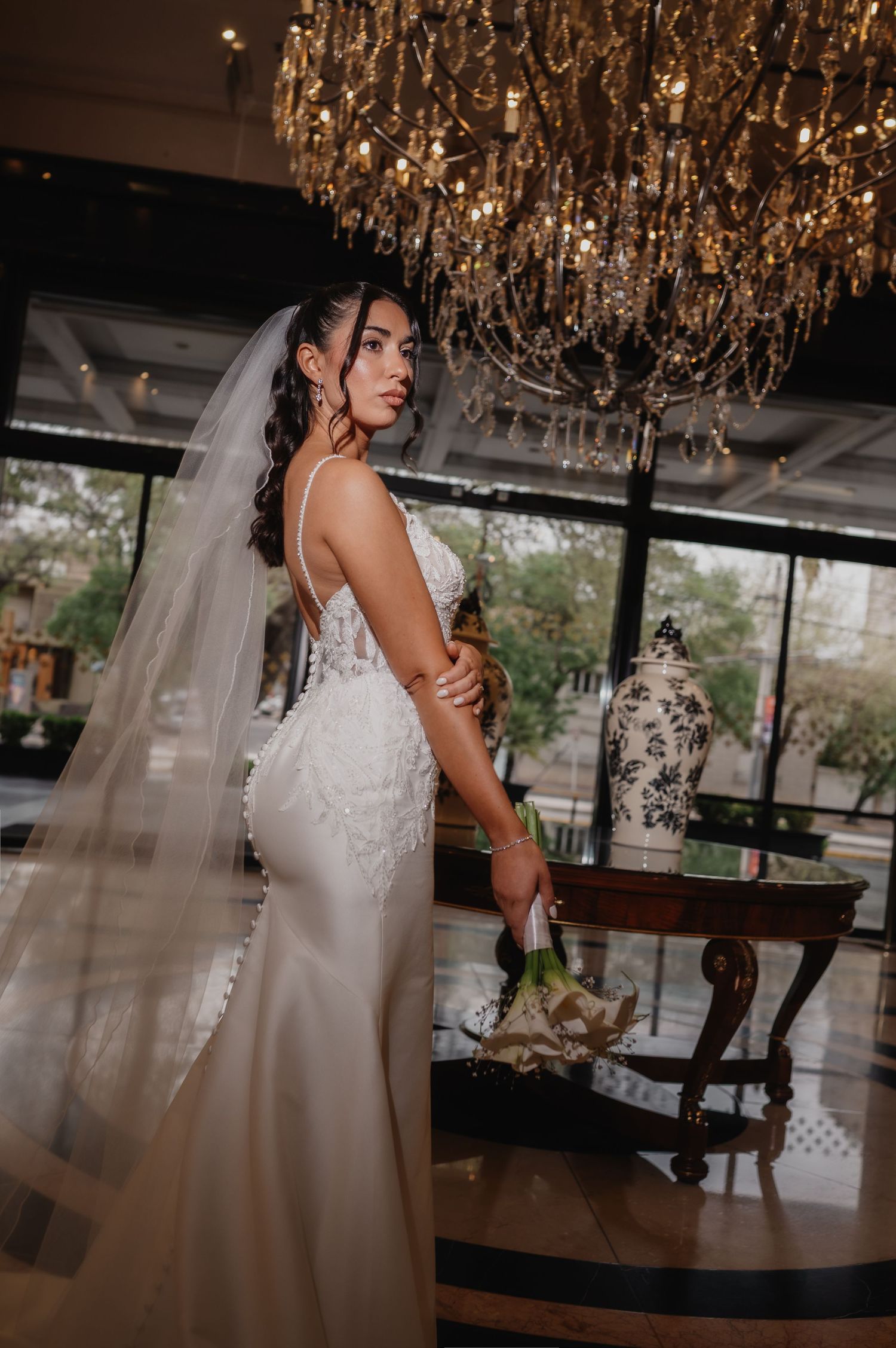 Elegant bride in lace wedding gown and veil holds bouquet under crystal chandelier in luxurious venue.
