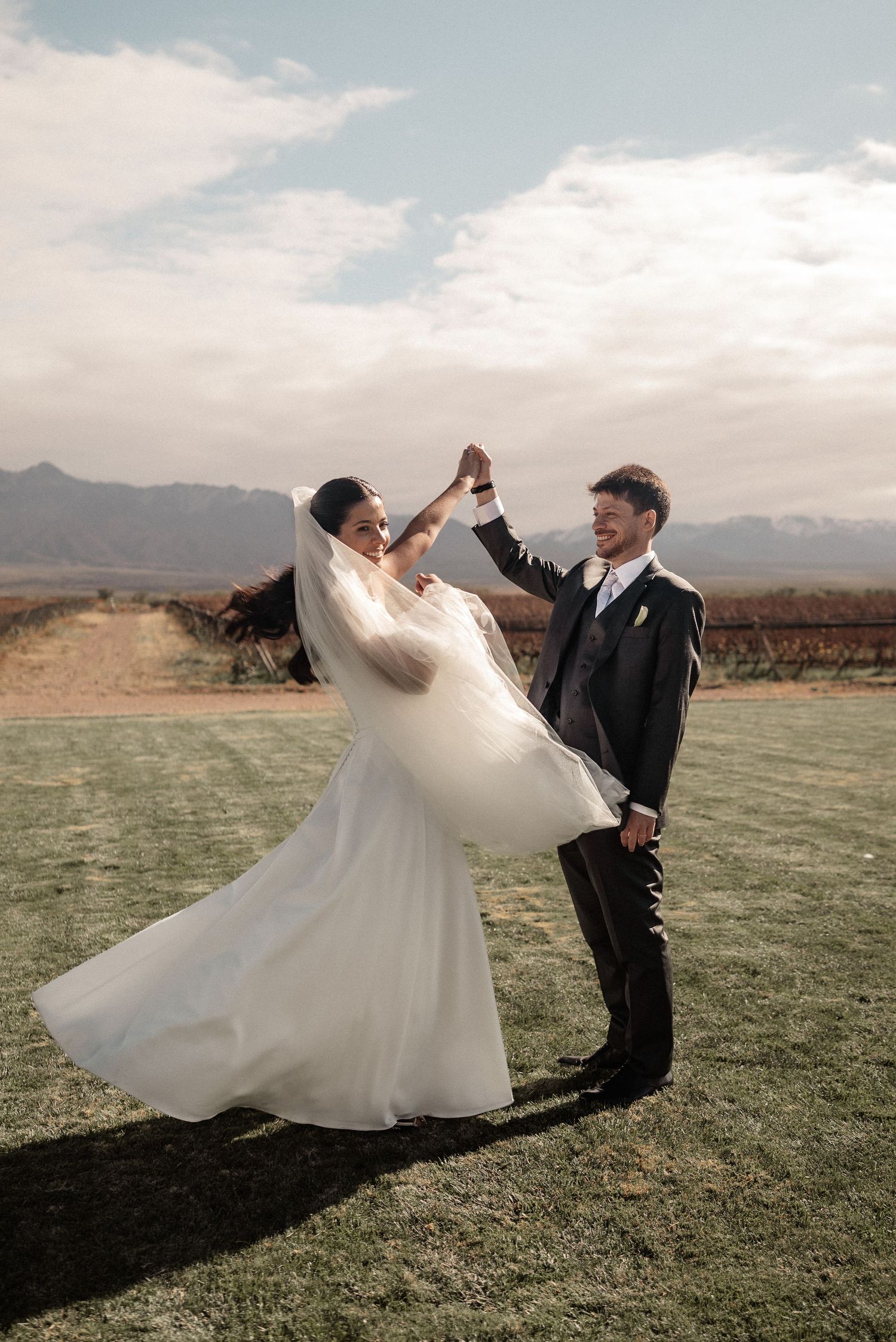 Bride and groom dancing outdoors at a vineyard wedding, with mountains and cloudy sky in the background.