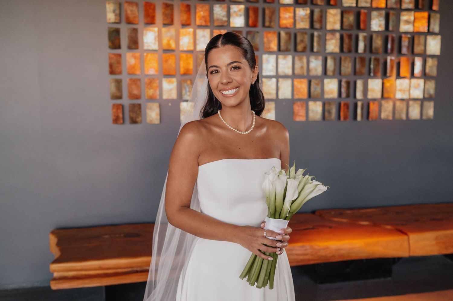 Smiling bride in strapless white gown holding white tulip bouquet with veil and pearl necklace indoors.