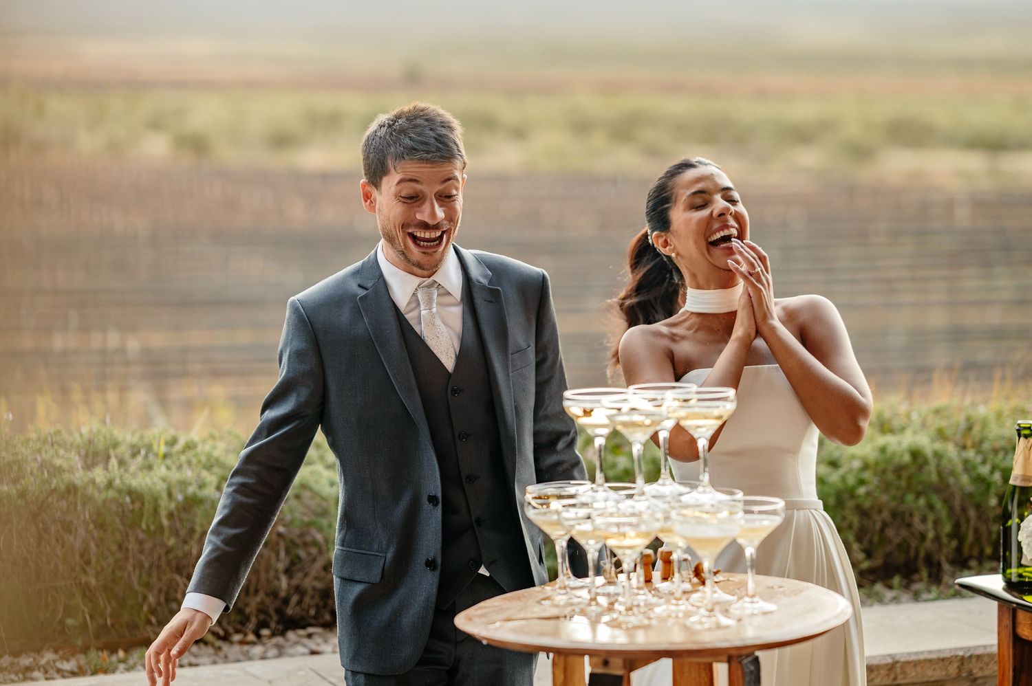 Joyful bride and groom laughing beside a champagne tower at an outdoor winery wedding reception.
