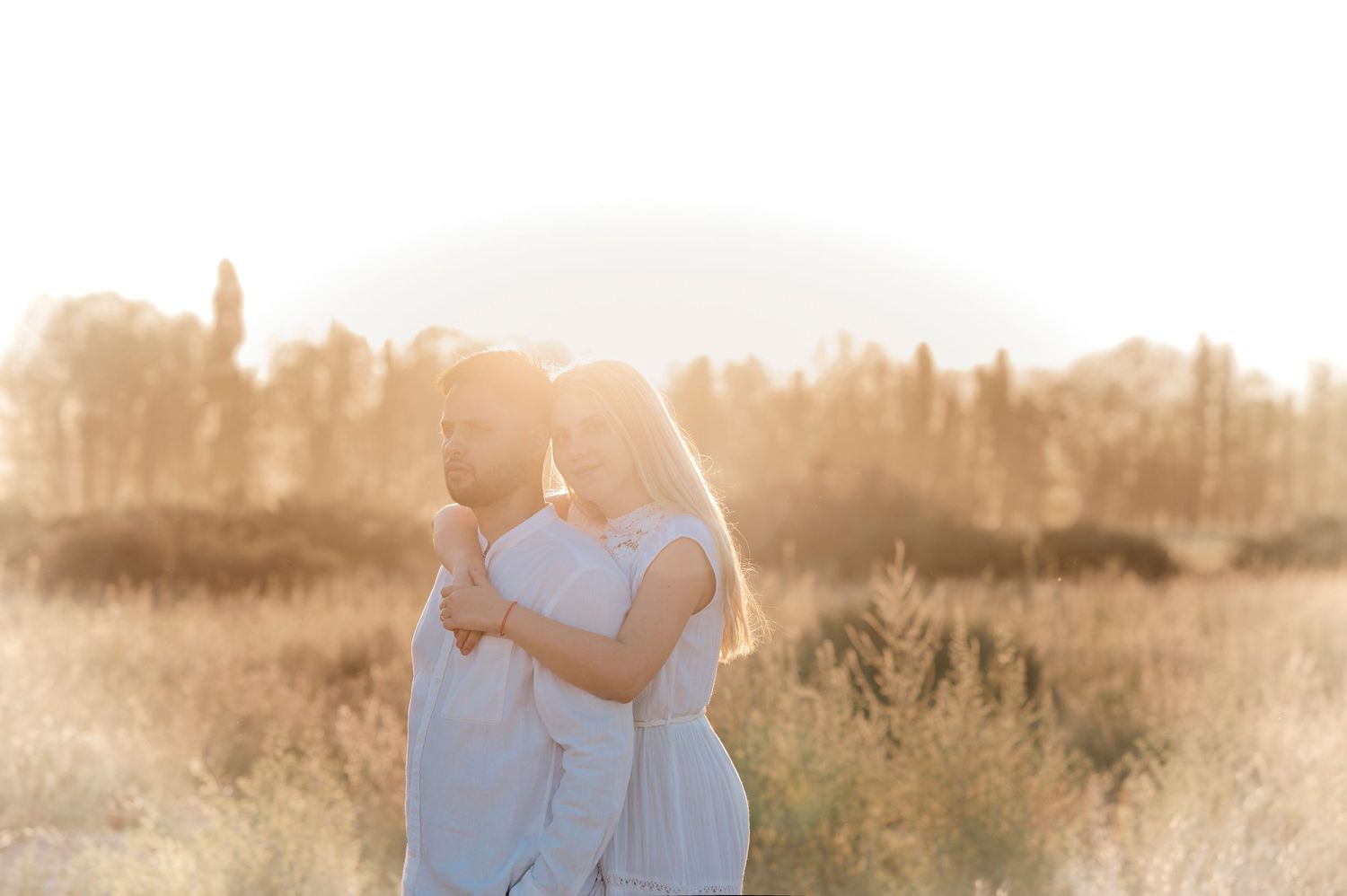 A couple stands together in a golden field at sunset creating a romantic silhouette against the warm sky.