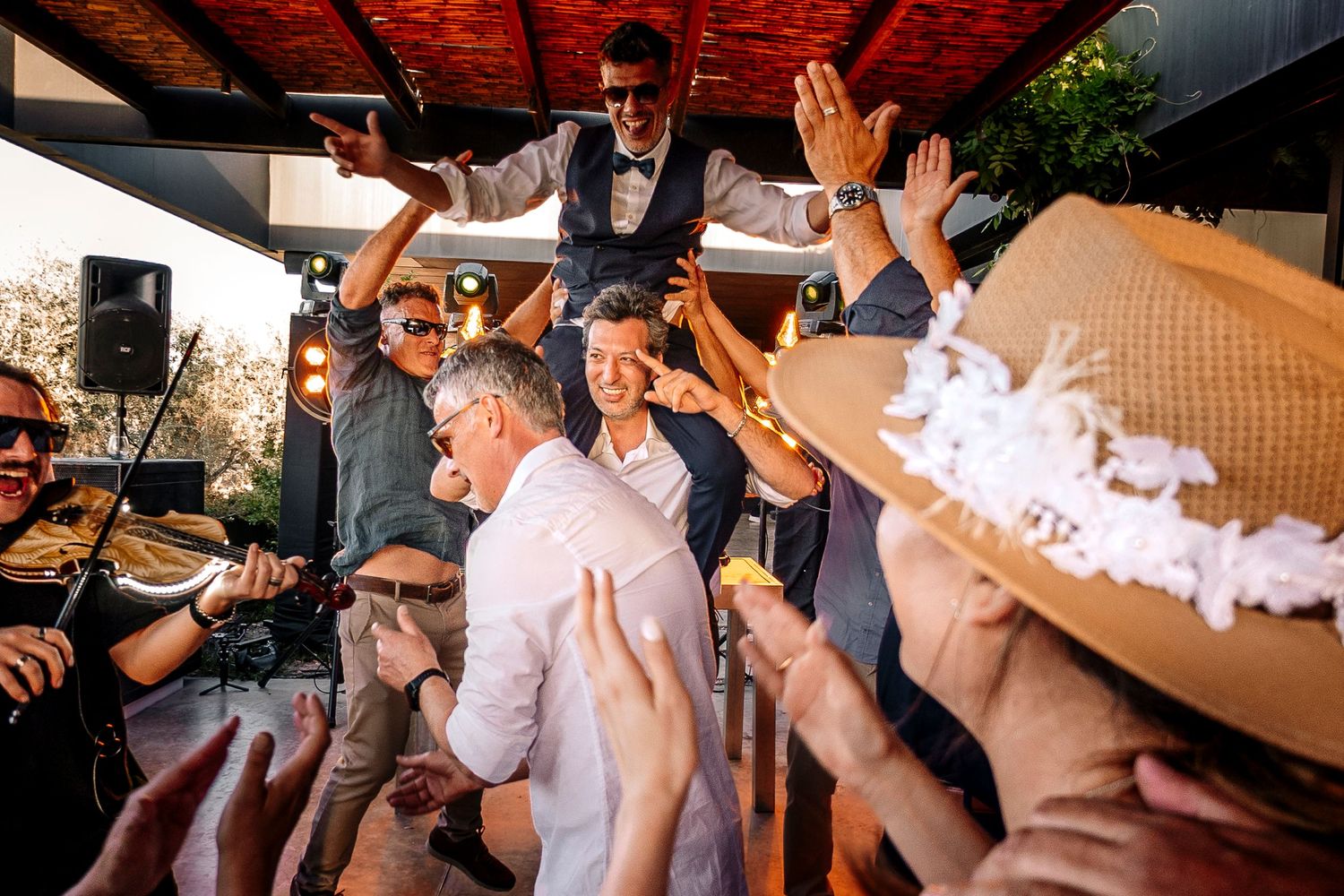 A lively crowd dances and celebrates at an outdoor party under a wooden pergola with hands raised in the air.