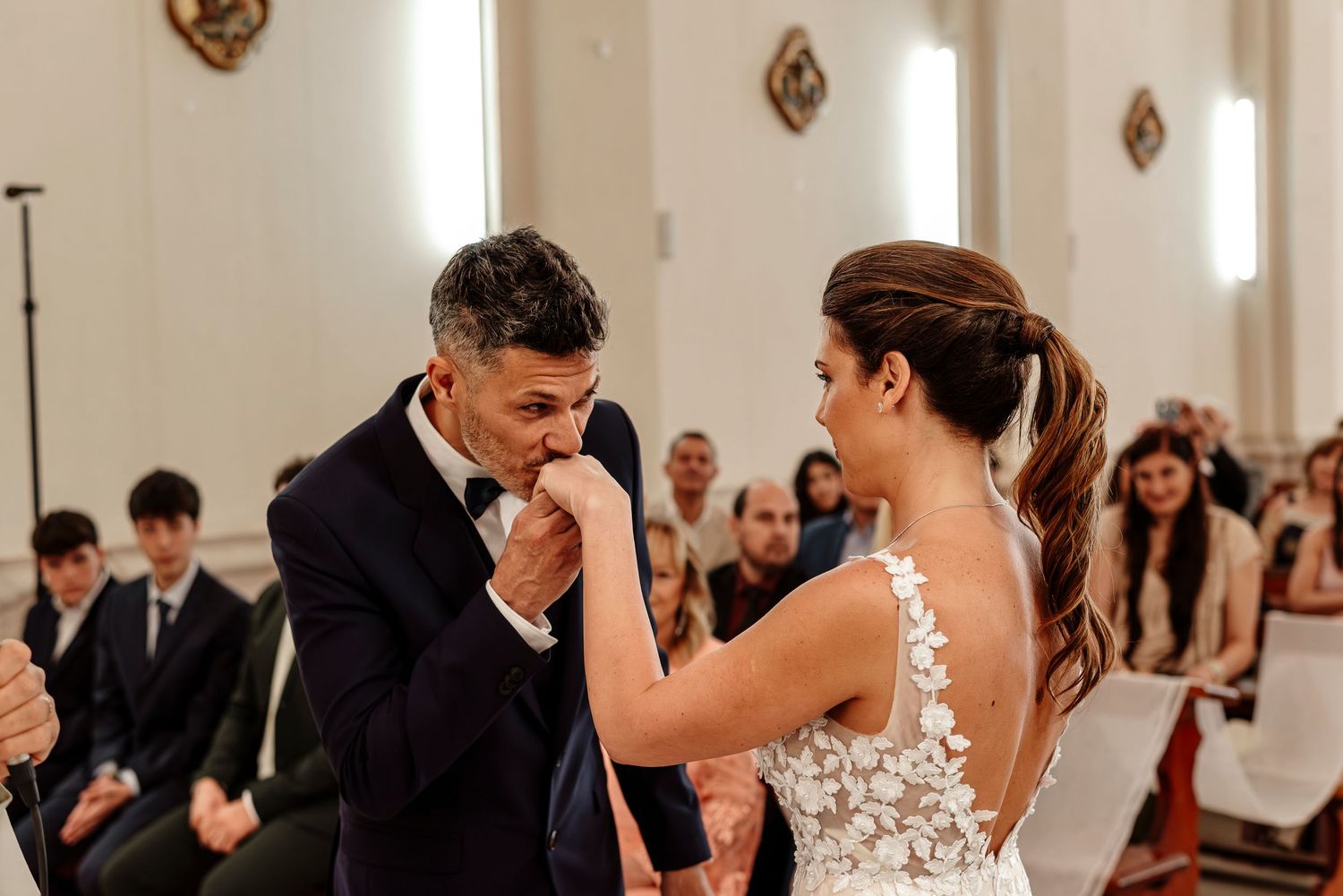 Wedding guests dance and celebrate during an elegant reception in a bright white ballroom with ornate wall decorations.