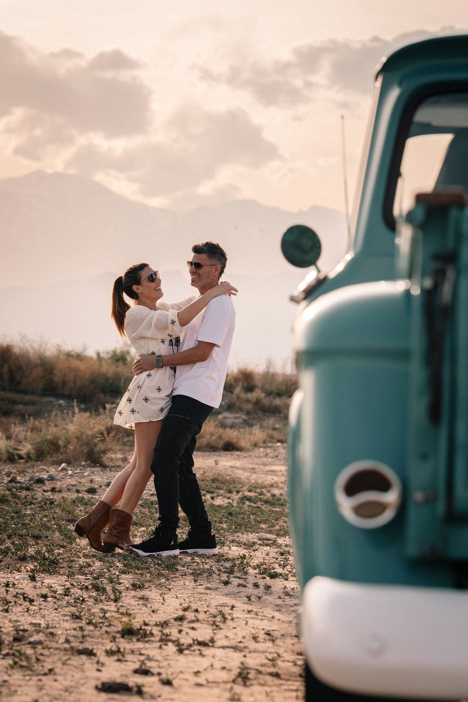 A couple embraces in a romantic desert photoshoot with a vintage turquoise vehicle in the background.