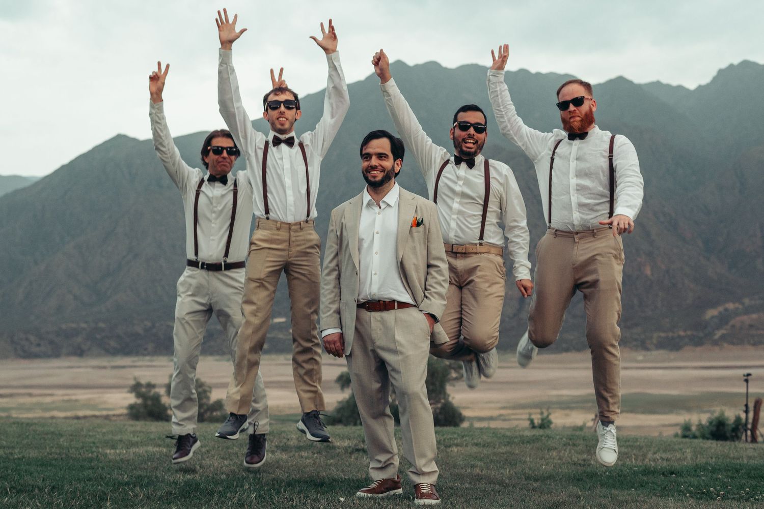Wedding party in light colored suits jumping together for a fun photo against a scenic mountain backdrop. fotografos de boda en mendoza