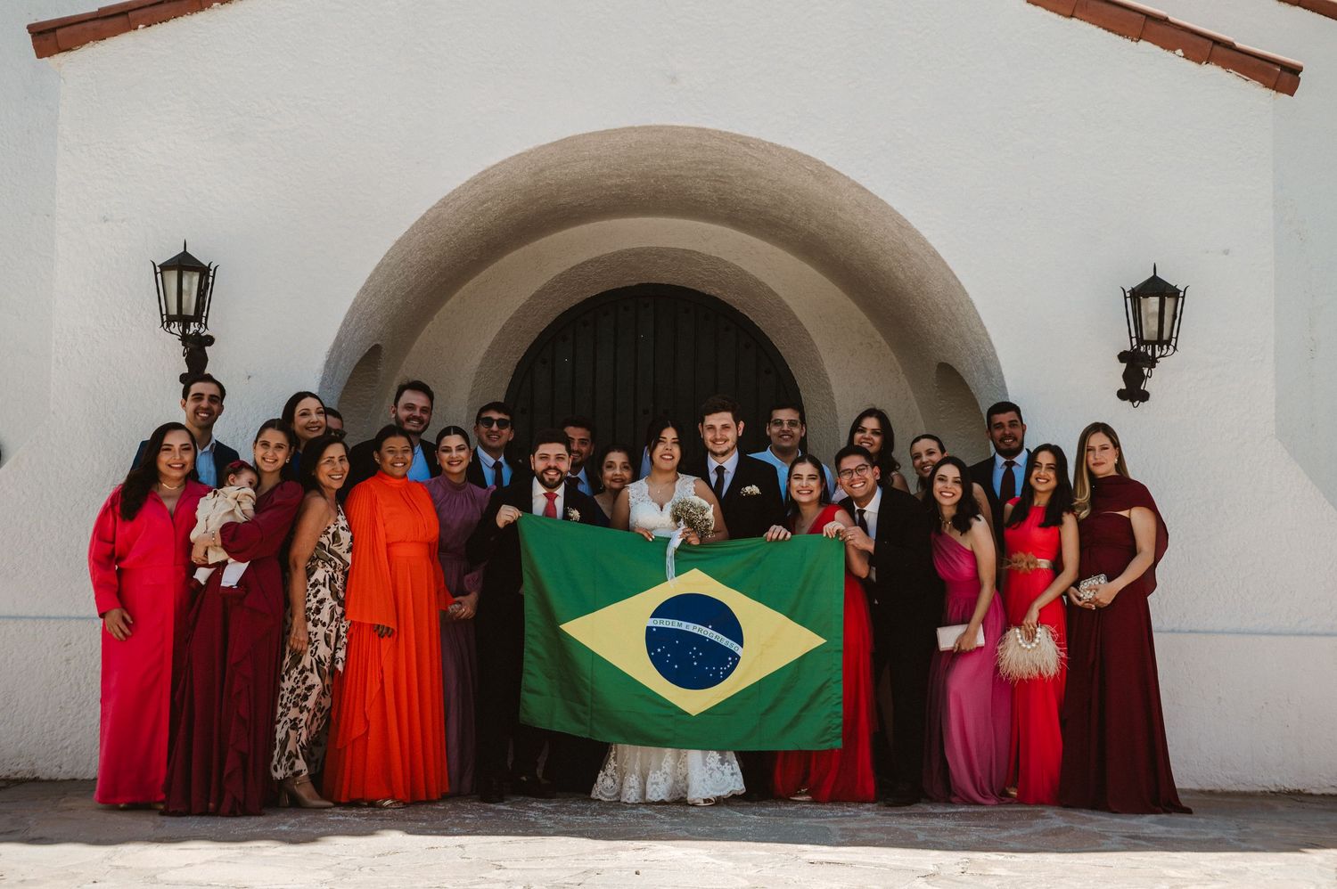 Wedding party guests in red and colorful formal dresses pose together for a group photo against white walls.