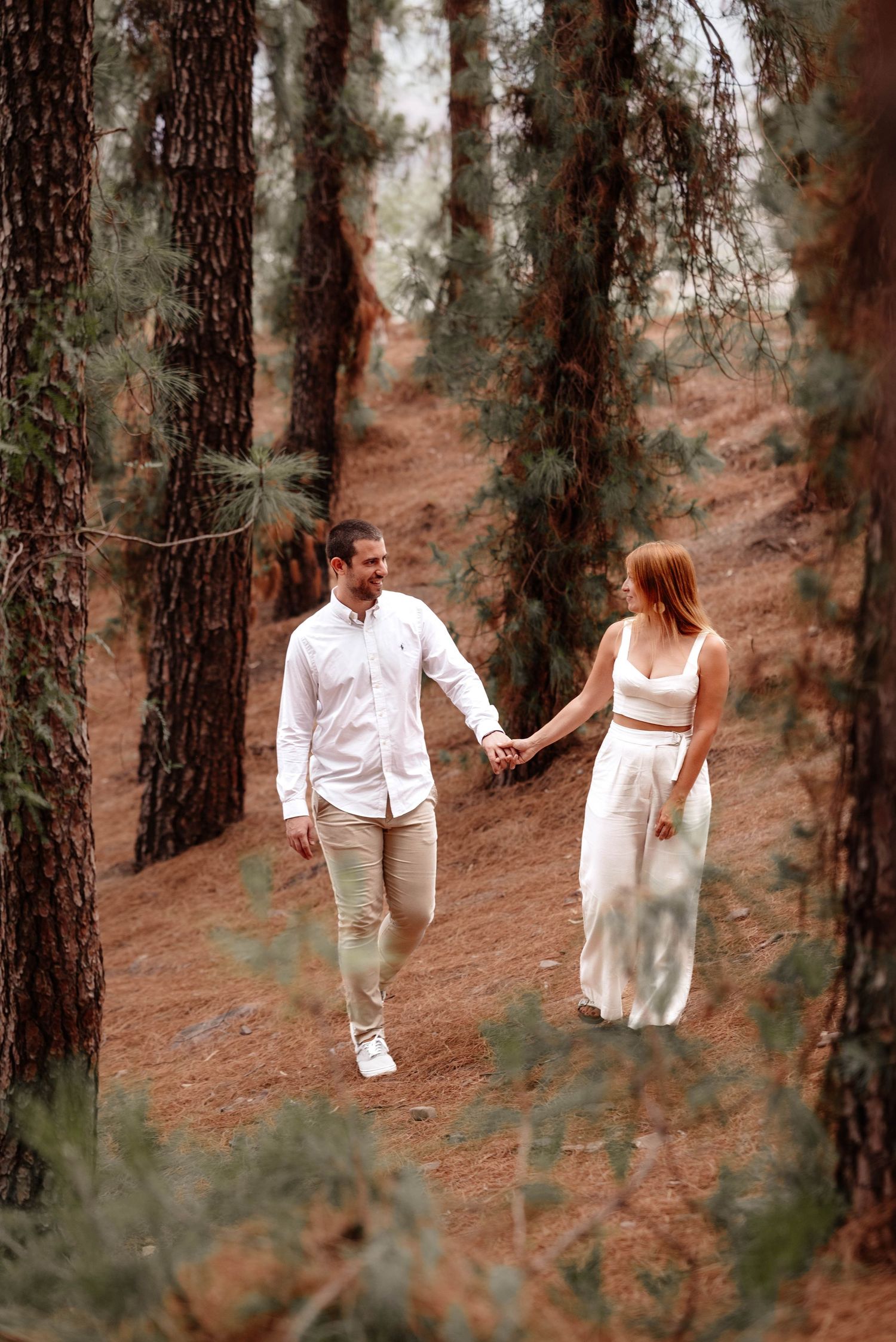 A couple in white outfits walks hand in hand through a pine forest on a romantic outdoor photo shoot.