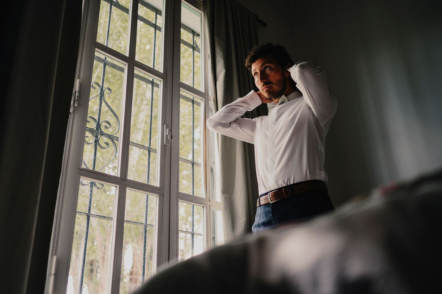 A person stands by large windows in a moody interior space with dramatic natural lighting and shadows.
