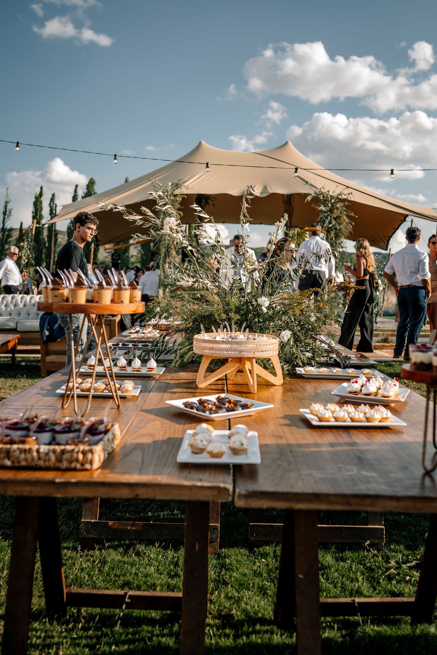 Outdoor catering setup with wooden tables displaying appetizers and desserts under tan canopies against a blue summer sky.