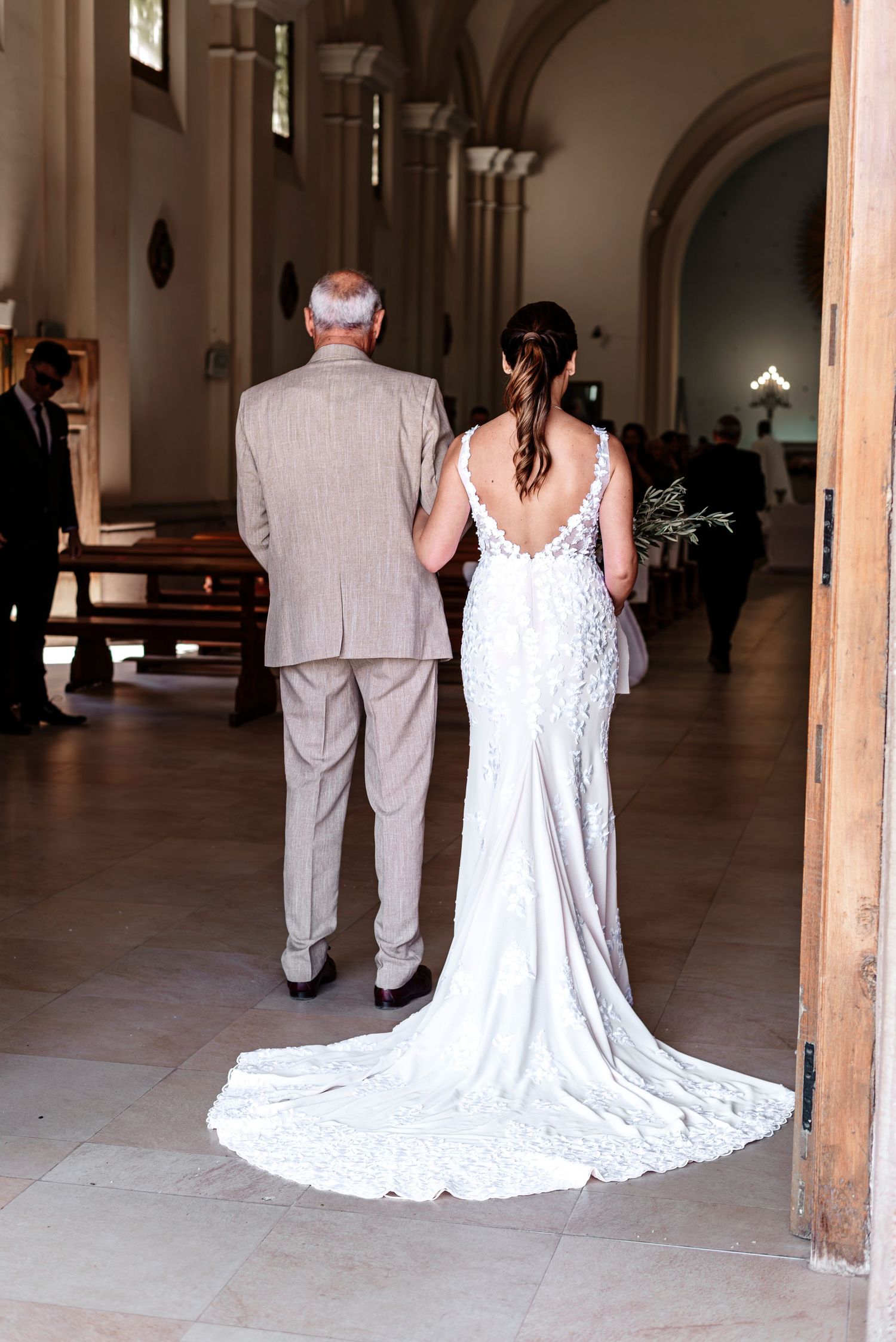 A wedding scene in an elegant hallway shows a bride in a white gown with a train following a person in a light suit.