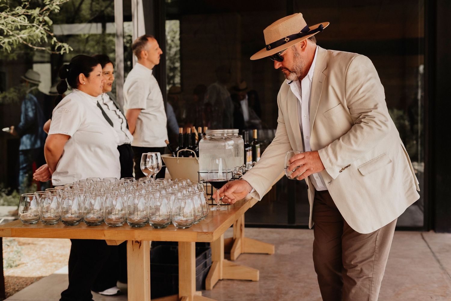 A person in a straw hat and beige jacket pours drinks at a rustic wooden table with rows of empty glasses.