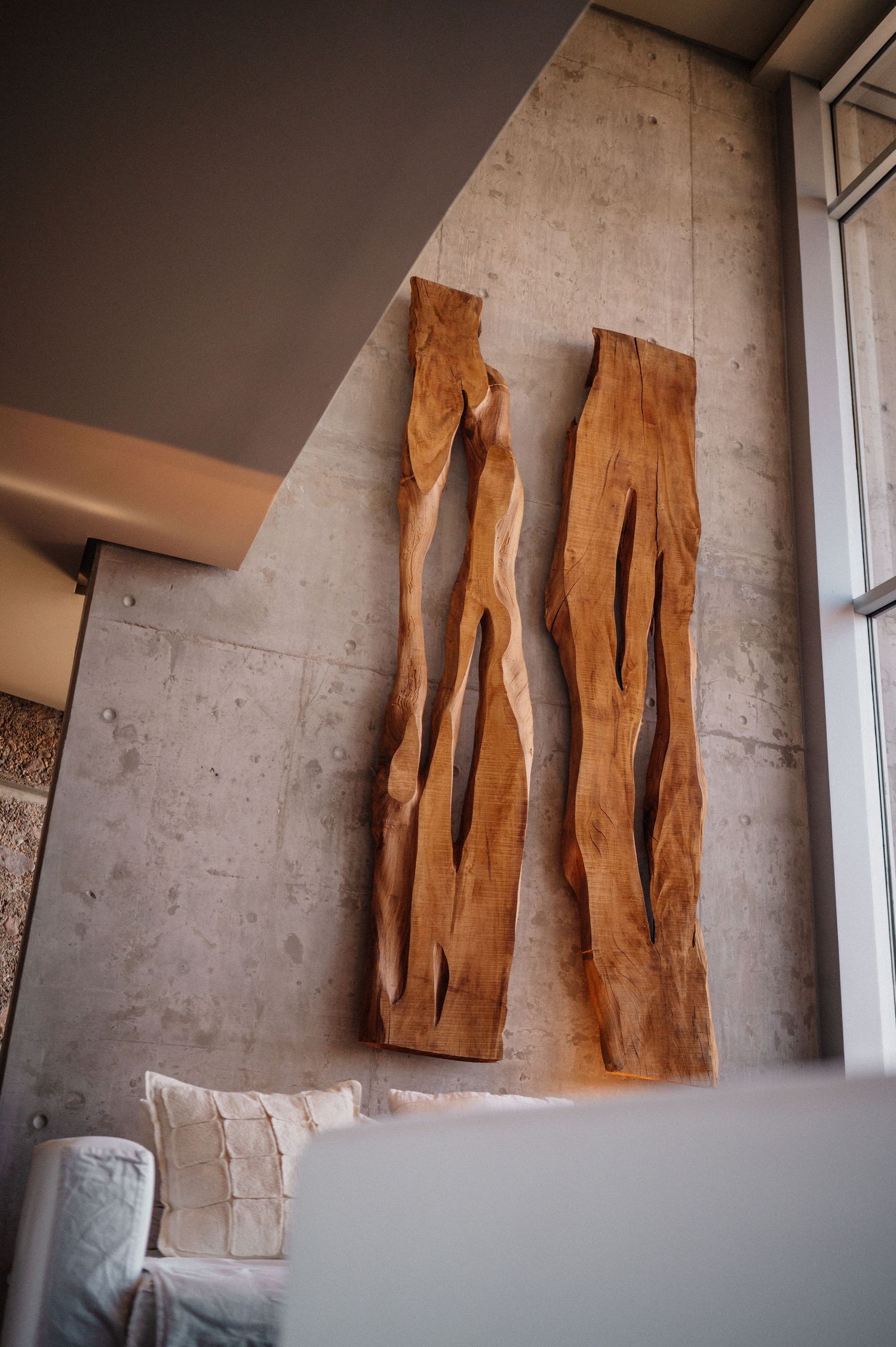 Two tall natural wood slab wall art pieces with organic cutouts mounted on a concrete wall above a white sofa.