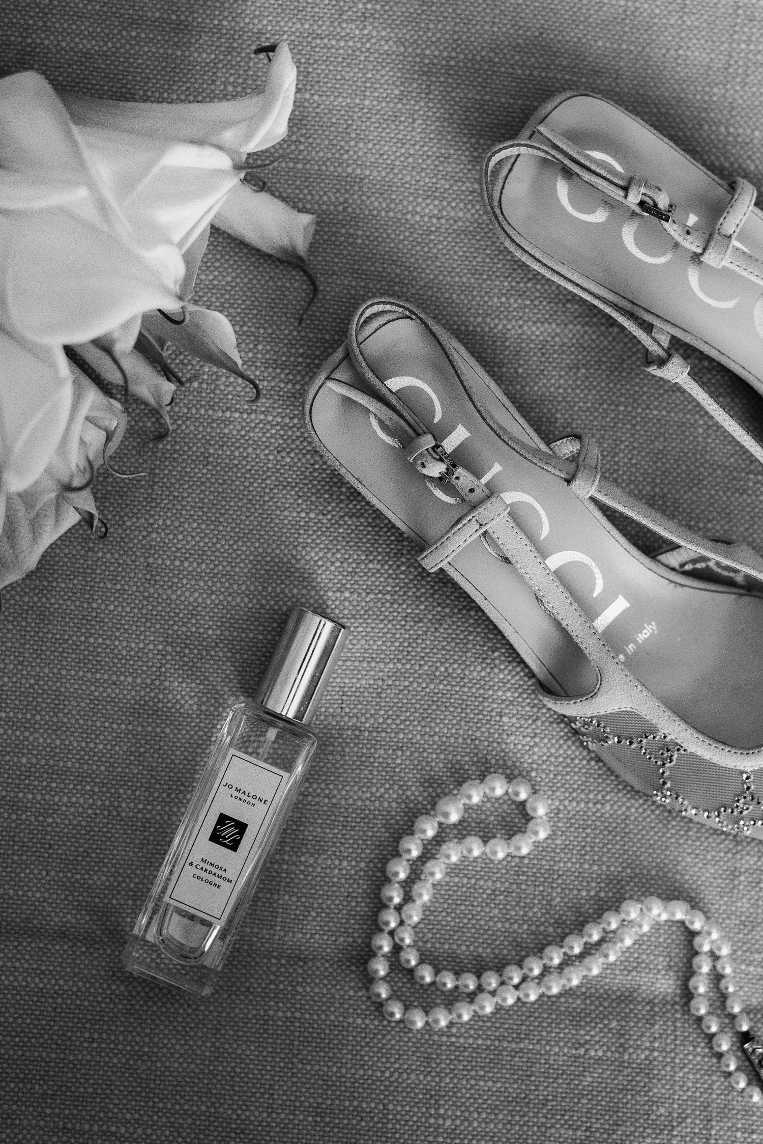 Black and white flatlay of Jo Malone perfume, pearl necklace, and Gucci loafers on textured fabric.