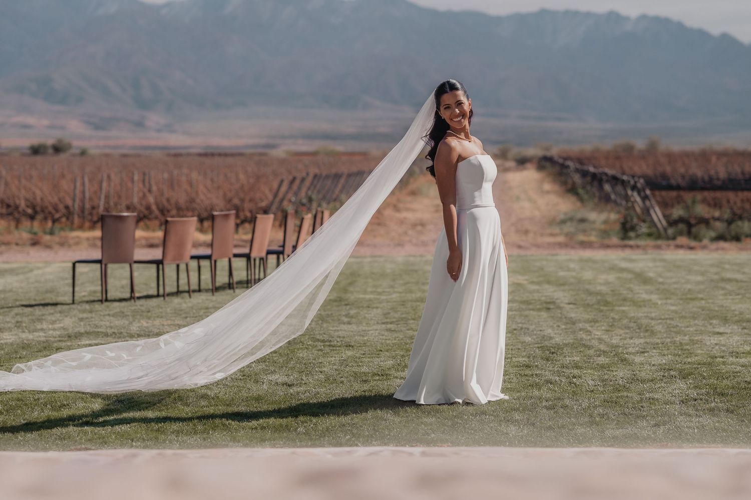 Bride in white gown with long veil at vineyard wedding venue, mountains in background.